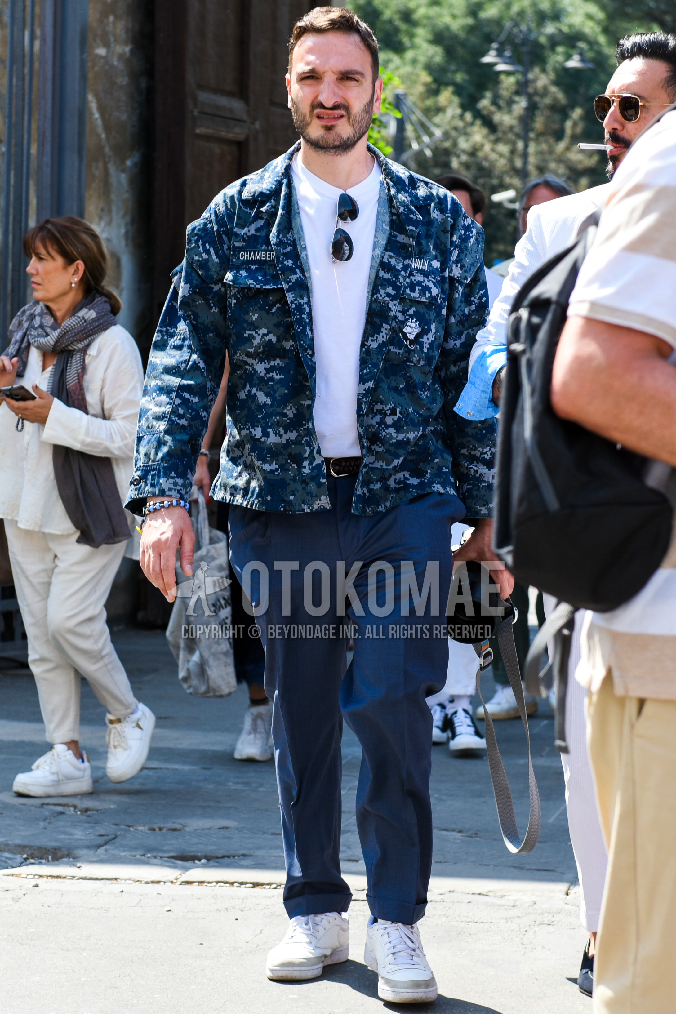 Men's spring summer autumn outfit with black plain sunglasses, navy camouflage military jacket, white plain t-shirt, navy plain chinos, white low-cut sneakers.