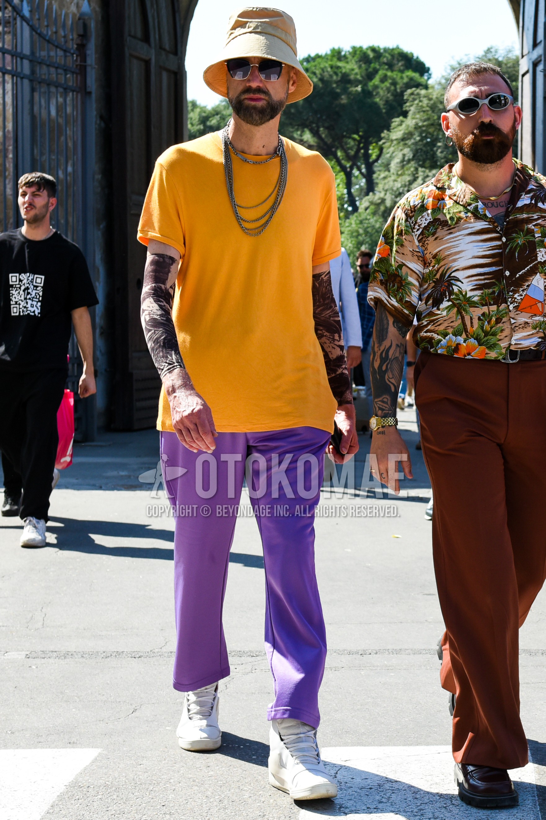 Men's spring summer outfit with beige plain bucket hat, gold plain sunglasses, orange yellow plain t-shirt, purple plain easy pants, purple plain ankle pants, white sneakers.