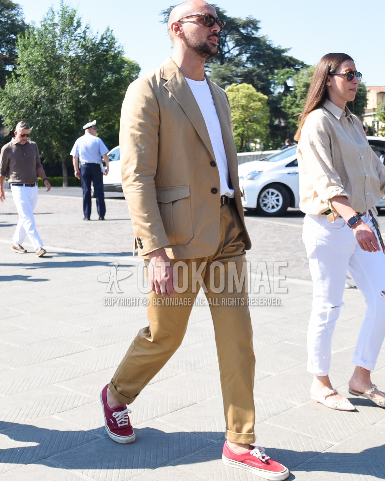 Men's spring summer outfit with brown tortoiseshell sunglasses, beige plain tailored jacket, white plain t-shirt, brown plain leather belt, beige plain chinos, red low-cut sneakers.