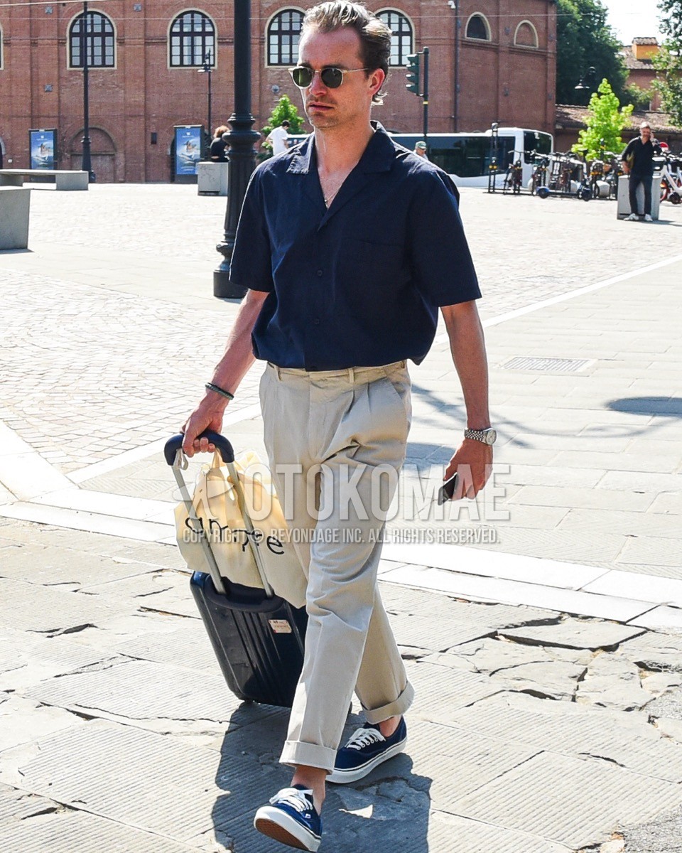 Men's spring summer outfit with clear plain sunglasses, navy plain shirt, white plain slacks, navy low-cut sneakers, black plain suitcase.