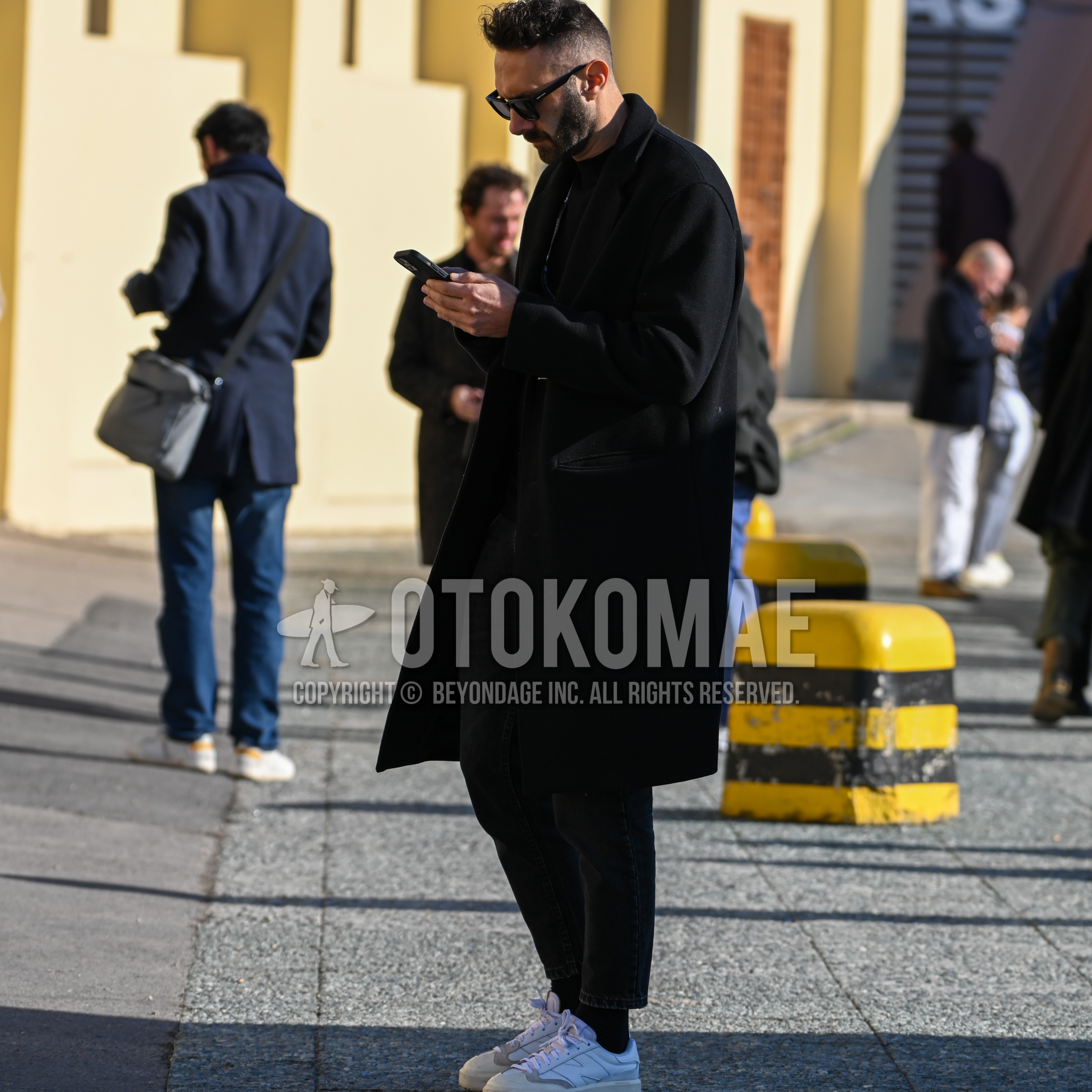 Men's autumn winter outfit with black plain sunglasses, black plain stenkarrer coat, black plain sweater, black plain ankle pants, black plain socks, white low-cut sneakers.