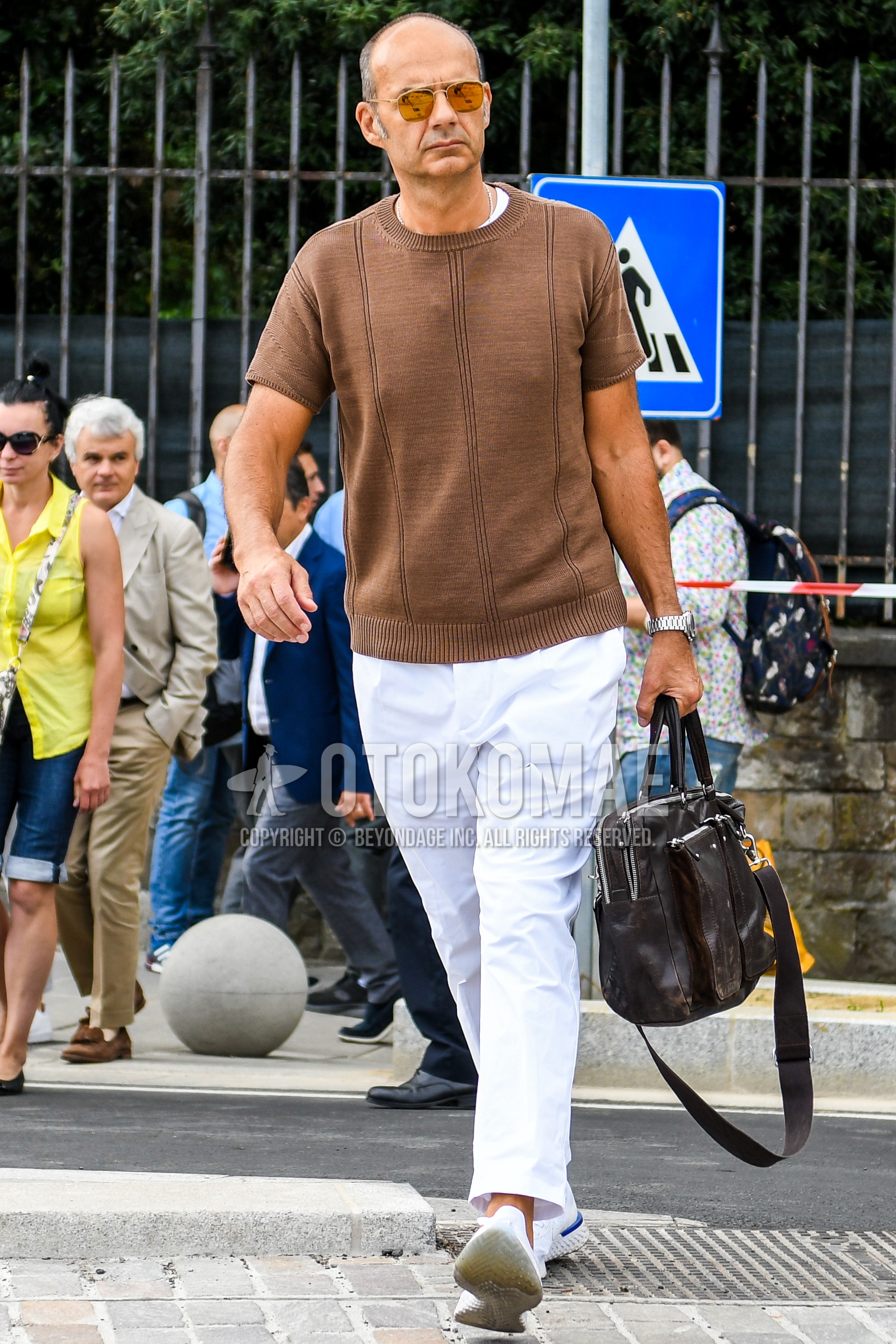 Men's summer outfit with plain sunglasses, beige plain sweater, white plain t-shirt, white plain ankle pants, white low-cut sneakers, brown plain briefcase/handbag.