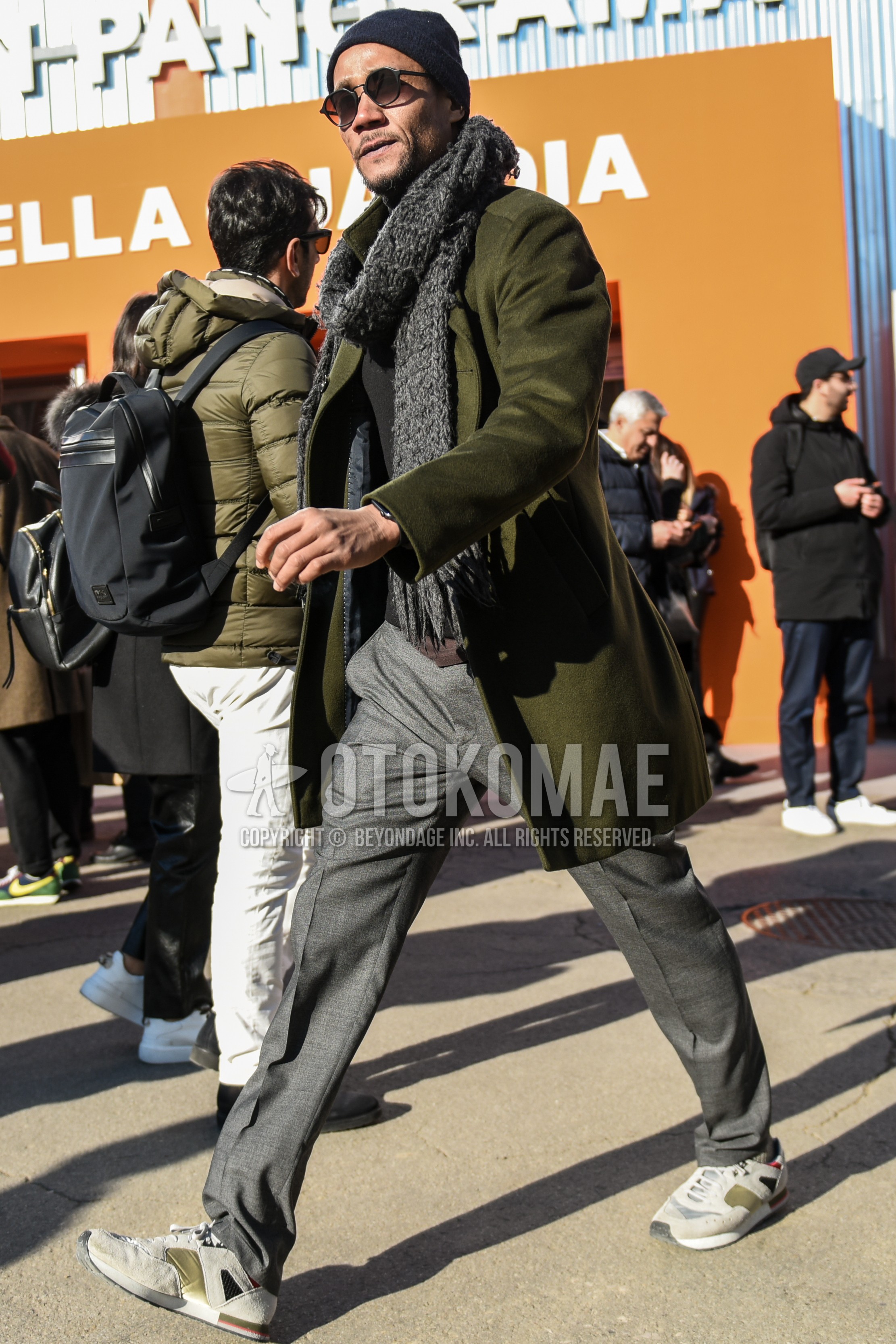 Men's autumn winter outfit with black plain knit cap, gray plain sunglasses, gray plain scarf, olive green plain stand collar coat, black plain sweater, gray plain slacks, gray low-cut sneakers.
