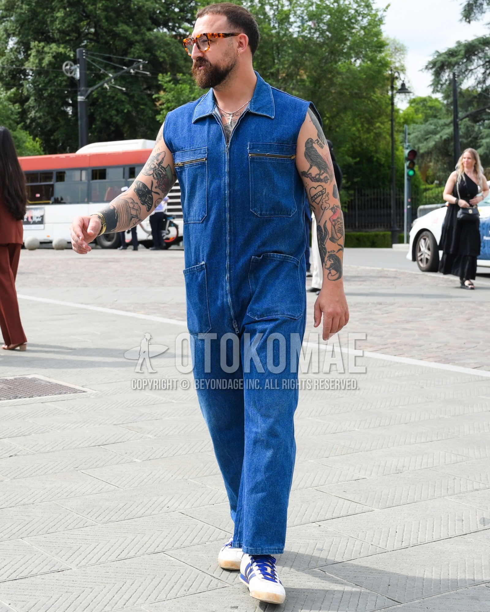 Men's spring summer outfit with clear tortoiseshell sunglasses, blue plain jumpsuit, white low-cut sneakers.