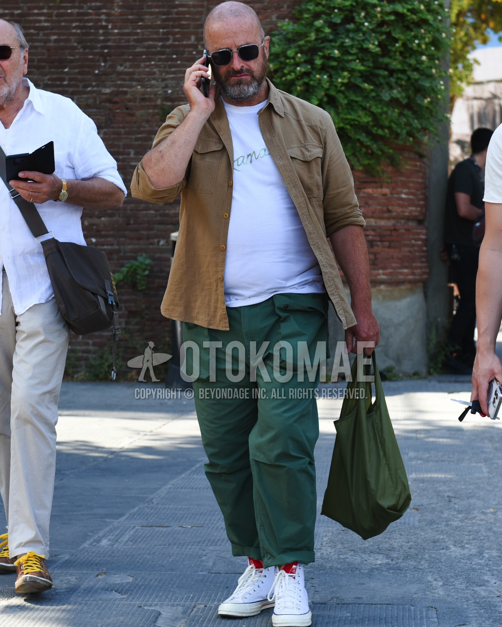 Men's spring summer outfit with silver plain sunglasses, brown plain shirt, white deca logo t-shirt, green plain cargo pants, white high-cut sneakers, olive green plain tote bag.