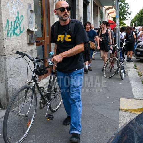 ウェリントンの黒無地のサングラス、黒レタードのTシャツ、ブルー無地のデニム/ジーンズ、黒スリッポンのスニーカー、黒スエードシューズの革靴、黒無地のバックパックを合わせた春夏のメンズコーデ・着こなし。