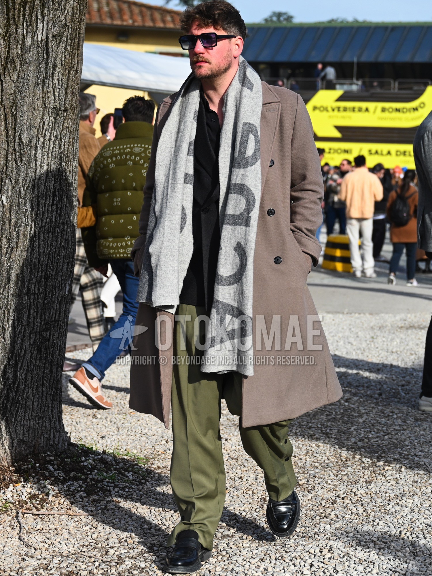Men's autumn winter outfit with black plain sunglasses, green deca logo scarf, brown plain ulster coat, black plain tailored jacket, black plain shirt, olive green plain slacks, black coin loafers leather shoes.