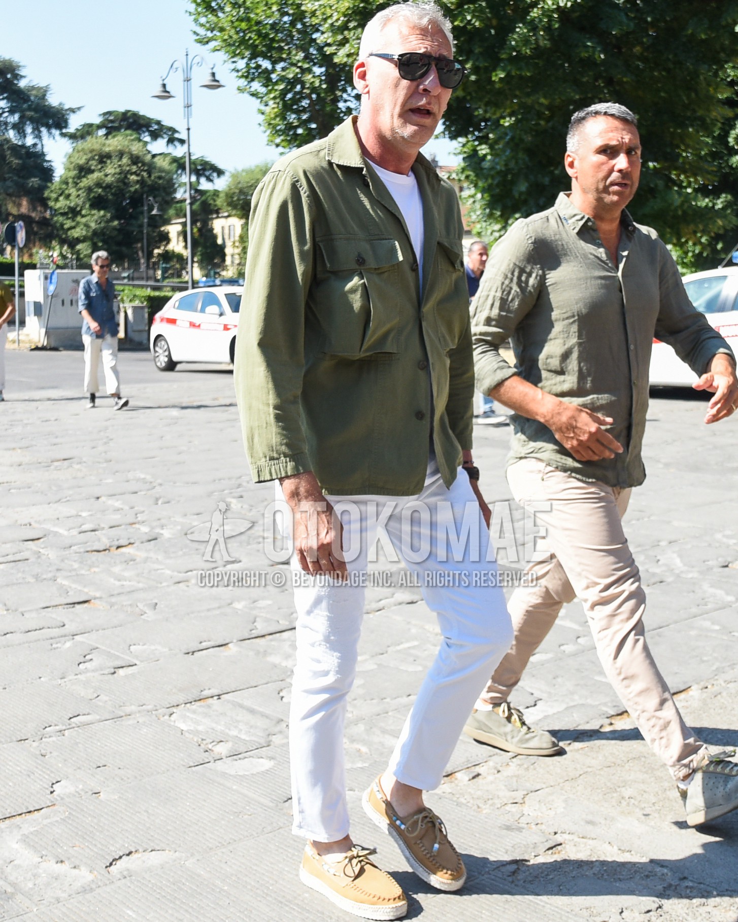 Men's spring summer outfit with black plain sunglasses, olive green plain field jacket/hunting jacket, white plain t-shirt, white plain cotton pants, beige moccasins/deck shoes leather shoes.