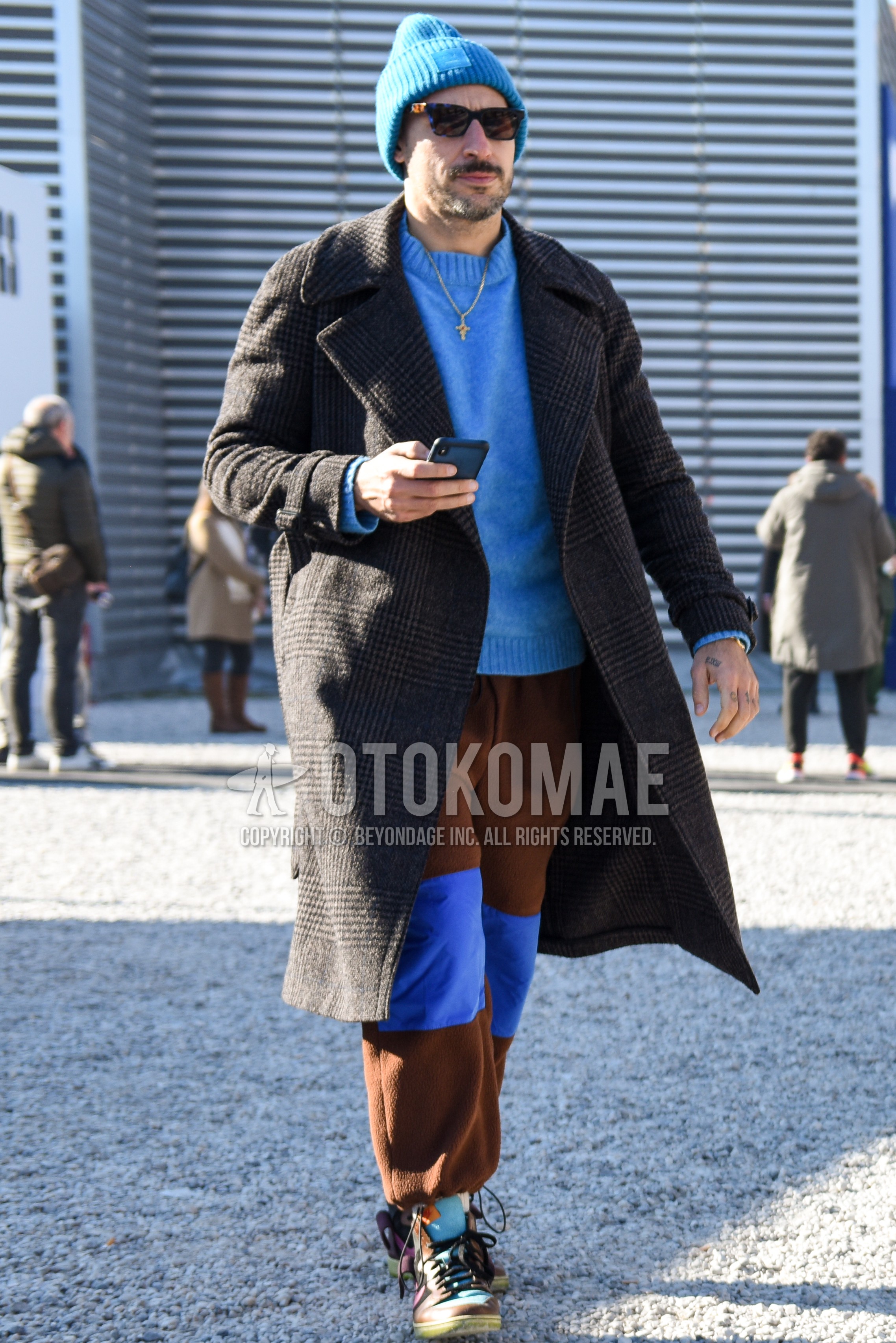 Men's autumn winter outfit with blue plain hat, blue tortoiseshell sunglasses, gray check trench coat, blue plain sweater, brown plain sweatpants, multi-color high-cut sneakers.