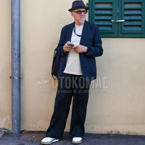 ネイビー無地のハット、黒無地のサングラス、ネイビー無地のテーラードジャケット、白無地のTシャツ、黒無地のスラックス、白ローカットのスニーカー、黒無地のバックパックを合わせた夏春のメンズコーデ・着こなし。