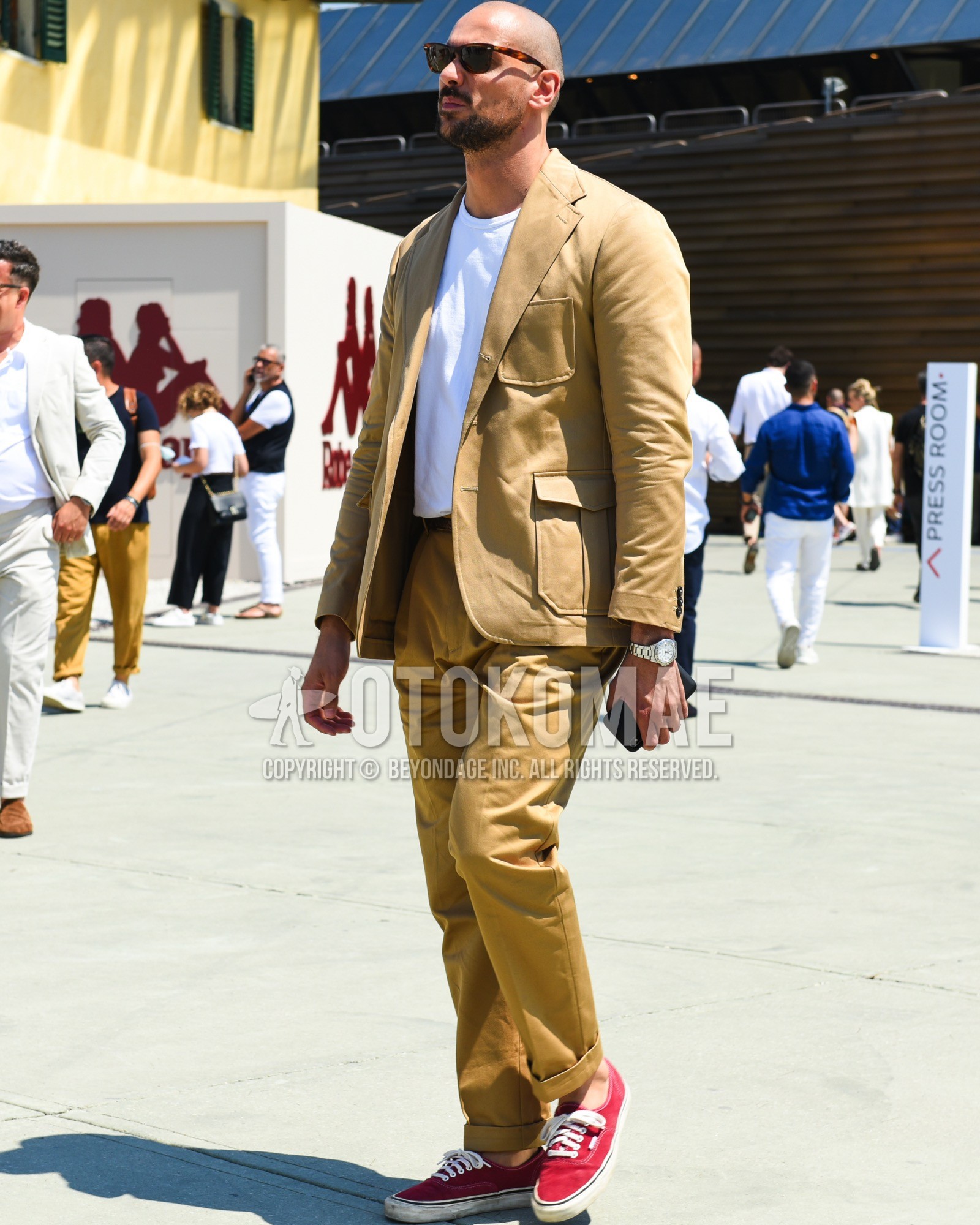 Men's spring summer outfit with brown tortoiseshell sunglasses, white plain t-shirt, brown plain leather belt, red low-cut sneakers, beige plain casual setup.