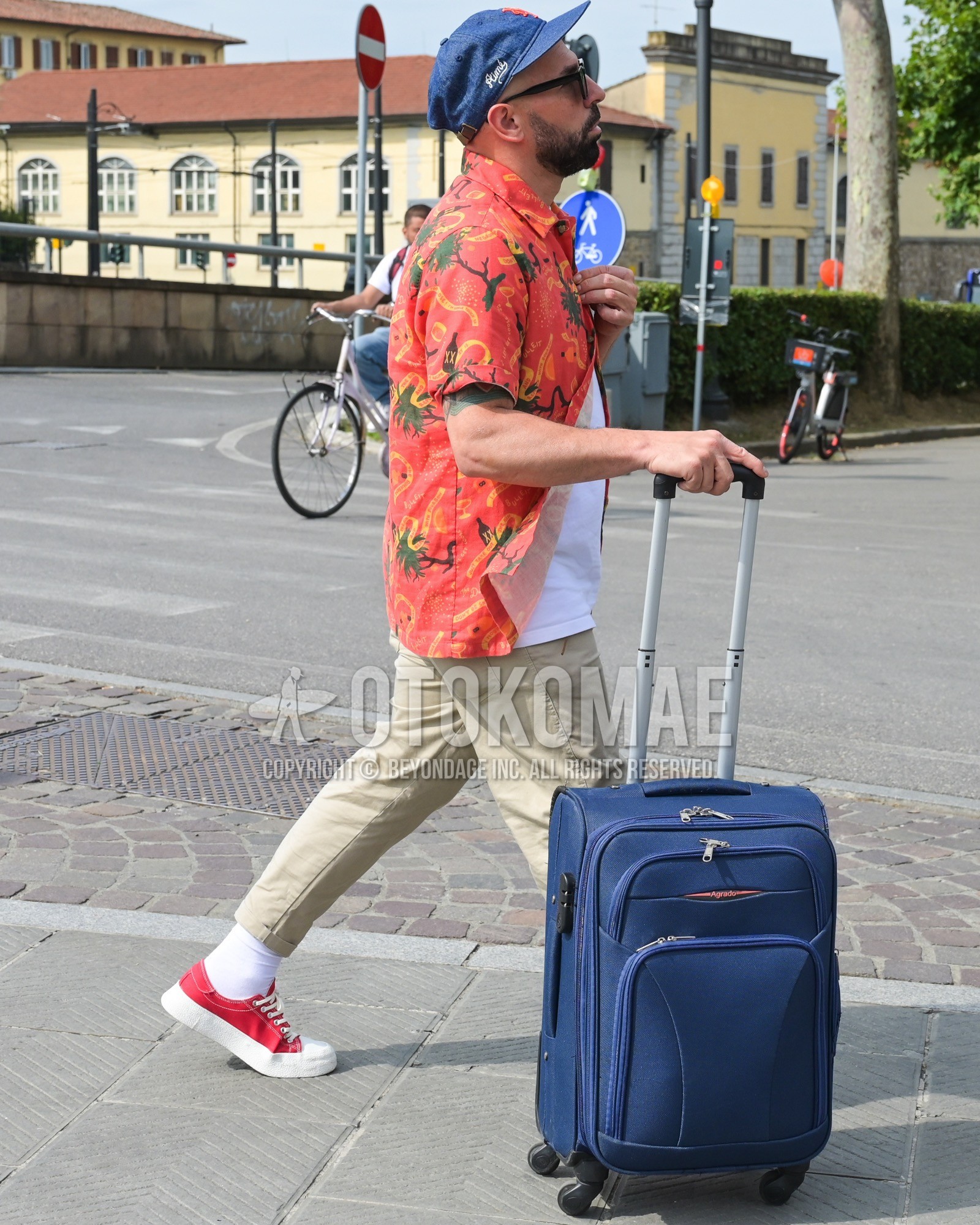 Men's spring summer outfit with blue one point baseball cap, orange whole pattern shirt, white plain t-shirt, beige plain chinos, white plain socks, red low-cut sneakers, navy plain suitcase.