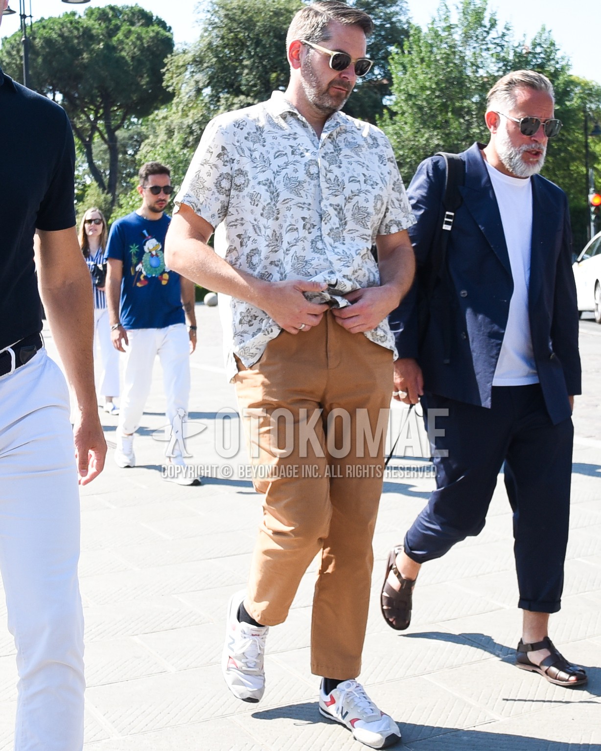 Men's spring summer outfit with beige plain sunglasses, white tops/innerwear shirt, brown plain chinos, black plain socks, white low-cut sneakers.