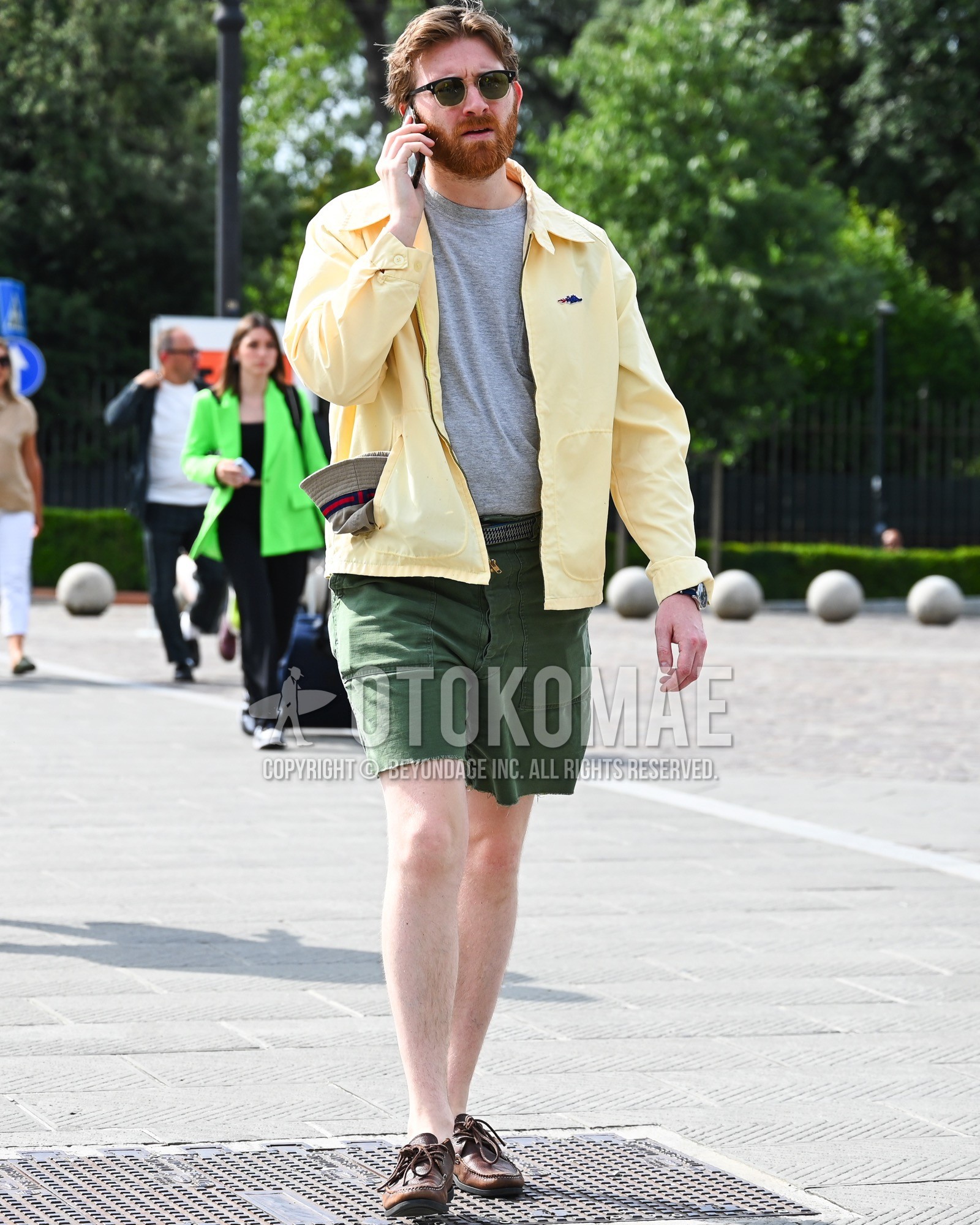 Men's spring summer outfit with black plain sunglasses, yellow one point shirt, green plain short pants, brown moccasins/deck shoes leather shoes.