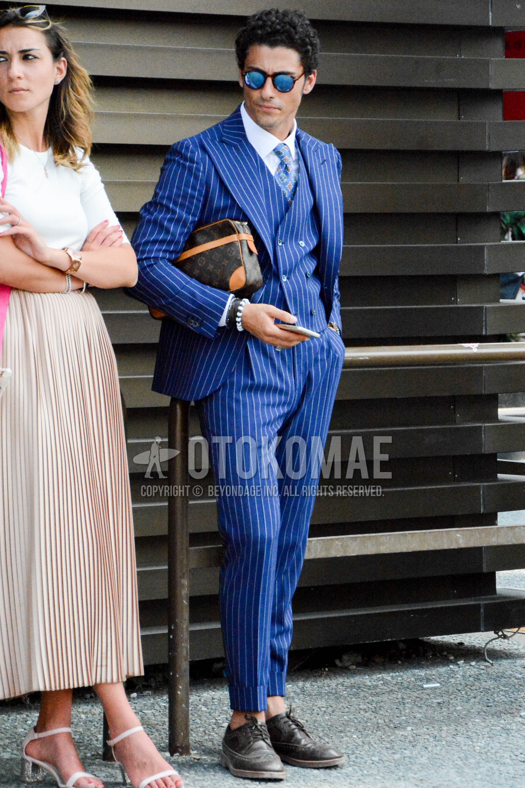 Men's spring autumn outfit with brown tortoiseshell sunglasses, white plain shirt, brown wing-tip shoes leather shoes, brown bag clutch bag/second bag/drawstring bag, navy stripes three-piece suit, blue small crest necktie.
