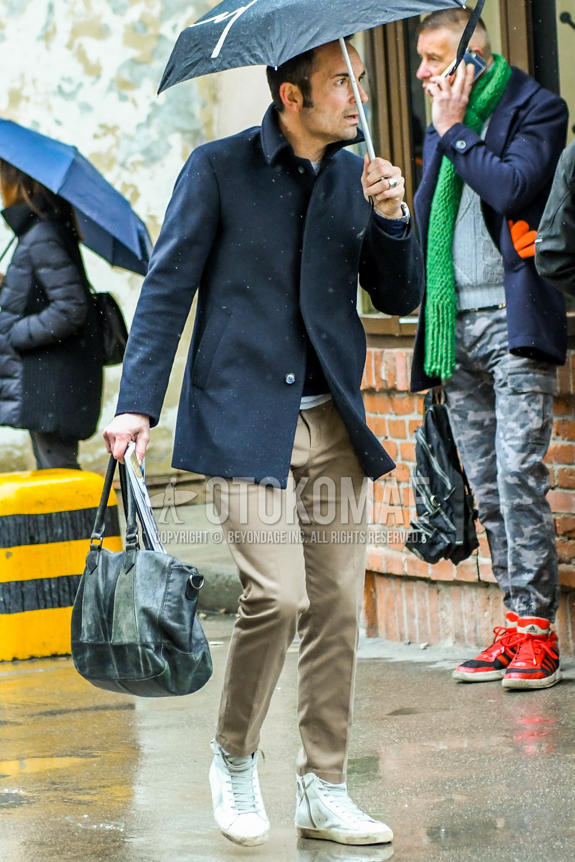 Men's autumn winter outfit with black plain stenkarrer coat, beige plain chinos, white high-cut sneakers, black plain briefcase/handbag.