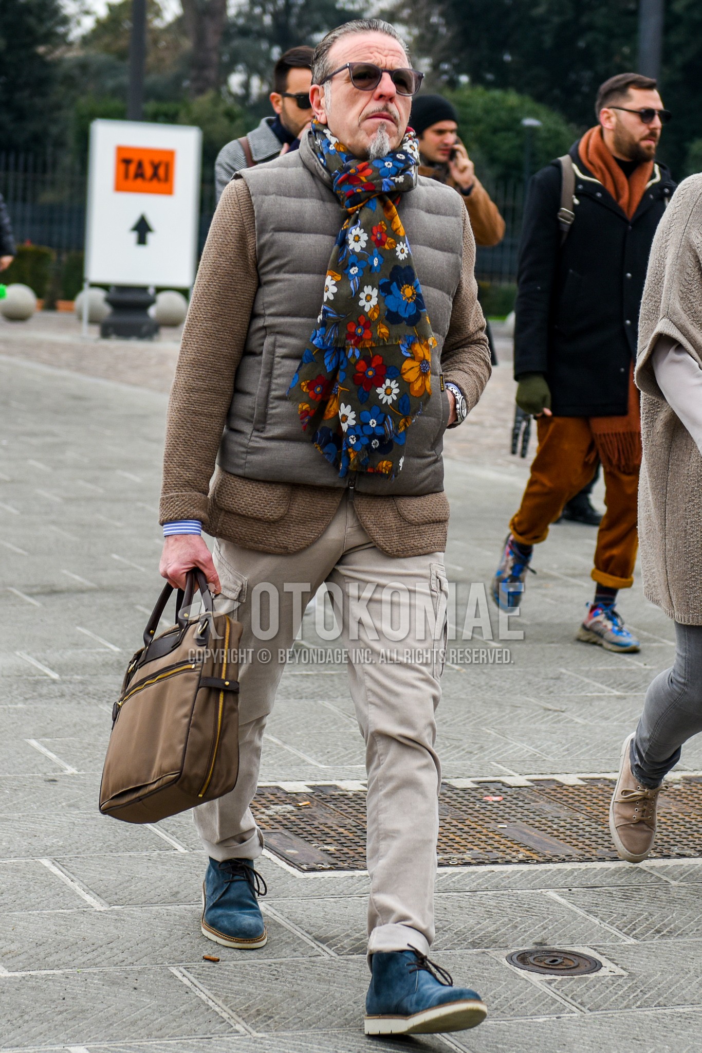 Men's winter outfit with plain sunglasses, multi-color botanical scarf, brown plain outerwear, brown plain tailored jacket, beige plain cargo pants, blue chukka boots, brown plain briefcase/handbag.