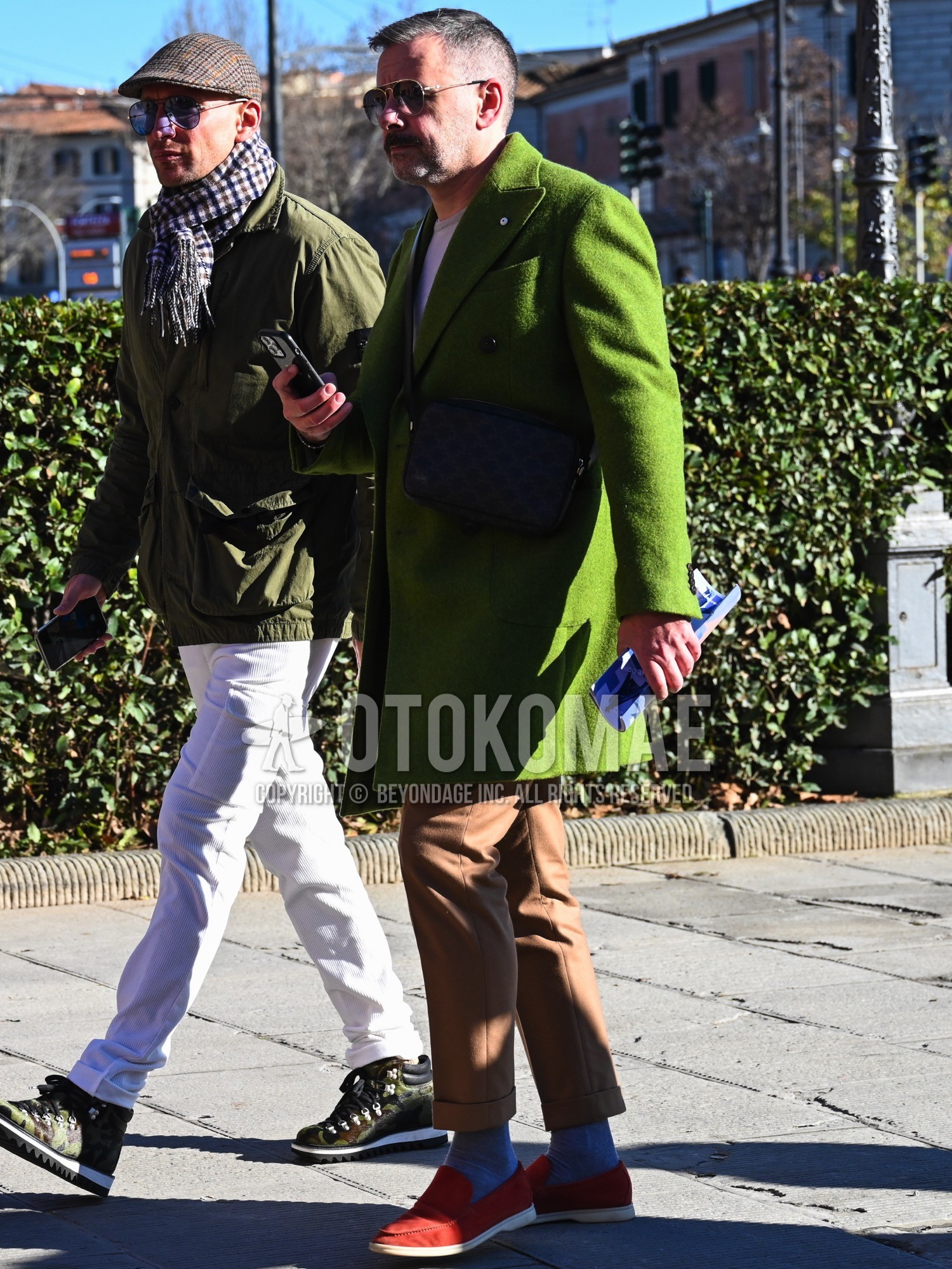 Men's autumn winter outfit with black plain sunglasses, green plain chester coat, gray plain long sleeve t-shirt, beige plain ankle pants, gray plain socks, red slip-on sneakers, black plain shoulder bag.