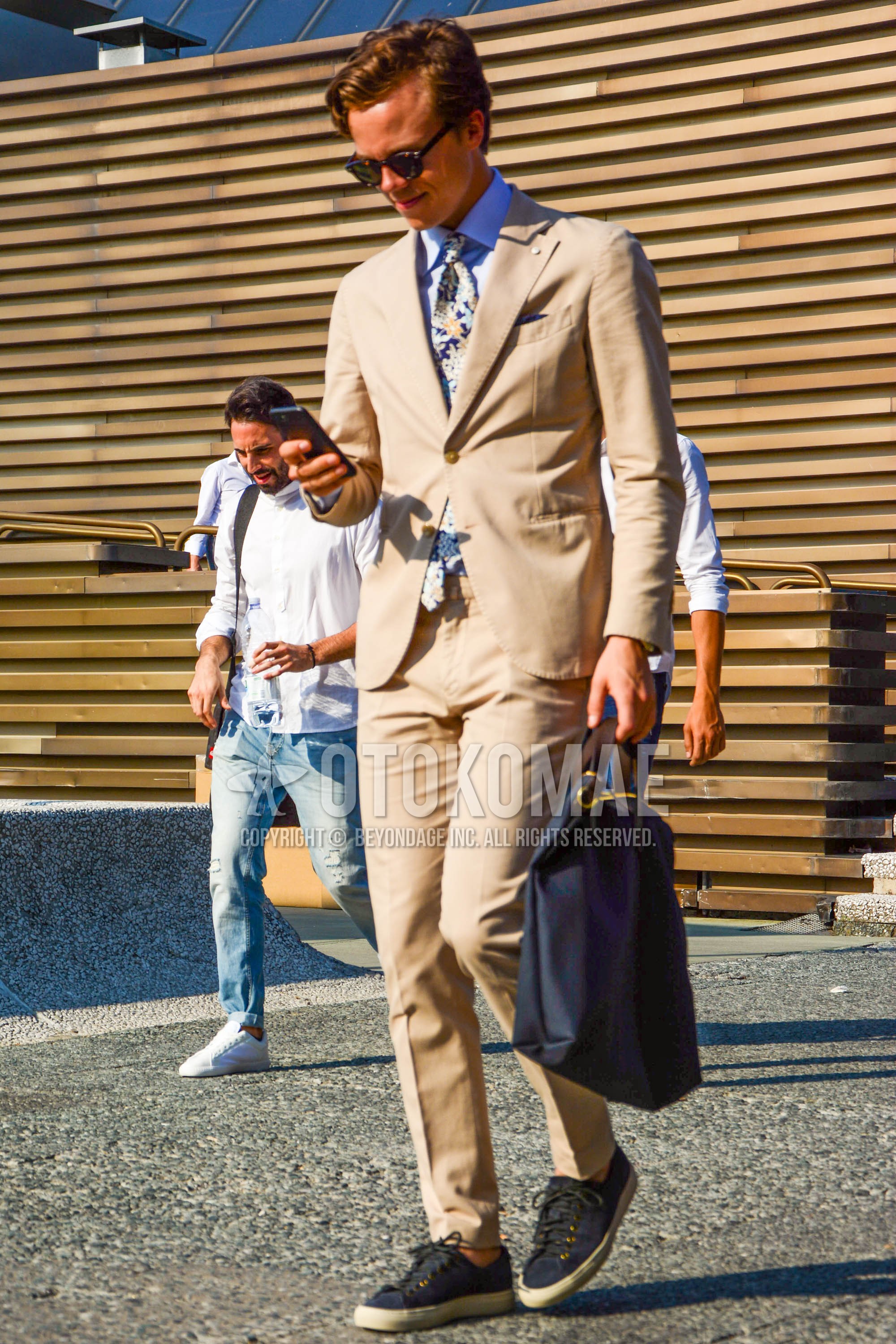 Men's spring autumn outfit with brown tortoiseshell sunglasses, light blue plain shirt, navy low-cut sneakers, black plain tote bag, beige plain suit, multi-color necktie necktie.