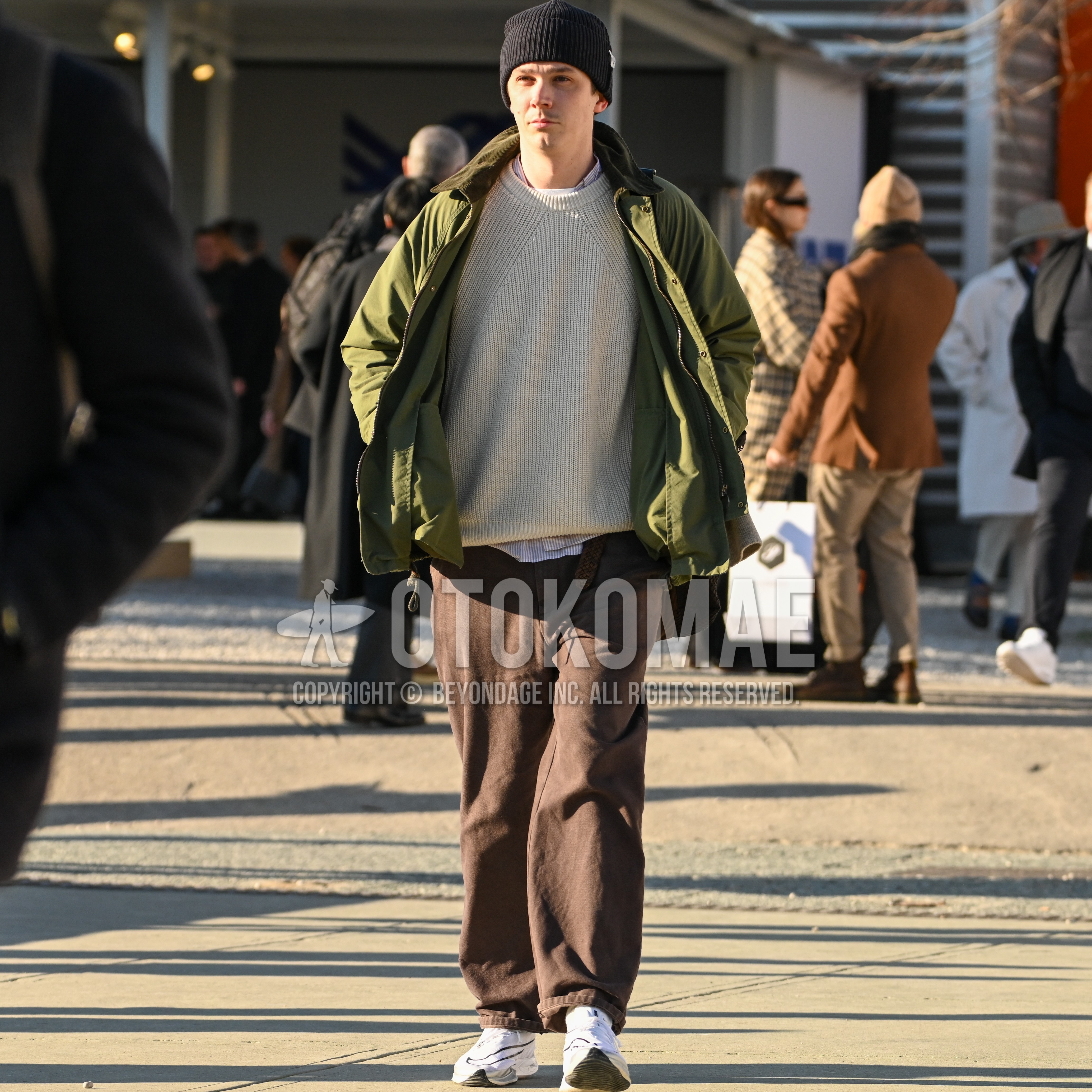 Men's autumn winter outfit with black one point knit cap, olive green outerwear field jacket/hunting jacket, stripes shirt, beige plain sweater, brown belt leather belt, brown plain denim/jeans, white low-cut sneakers.