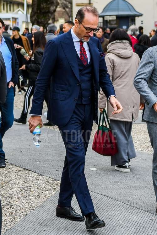 無地のメガネ、白無地のシャツ、黒ウィングチップの革靴、ネイビー無地のスリーピーススーツ、レッド・黒ネクタイのネクタイを合わせた春秋のメンズコーデ・着こなし。