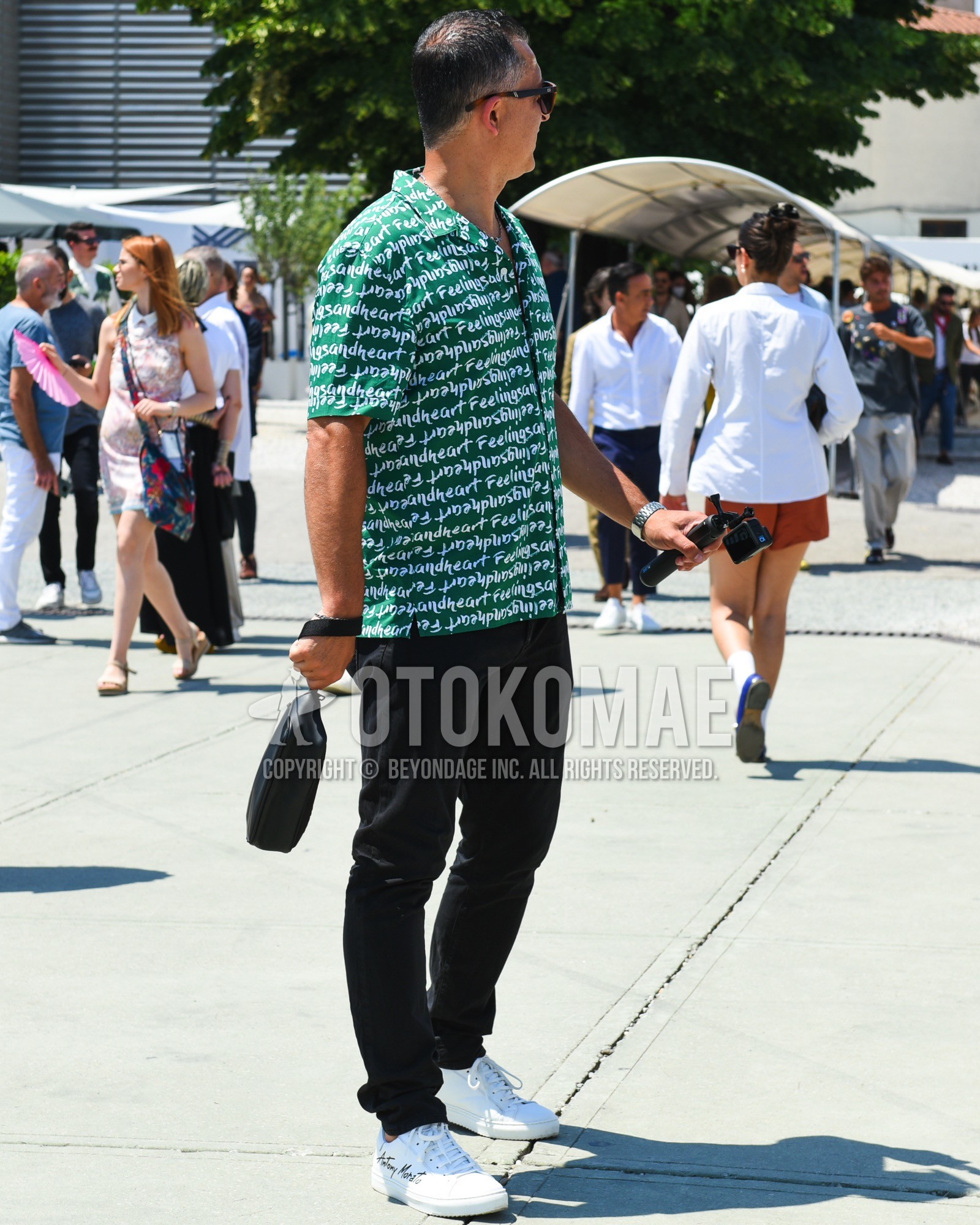 Men's spring summer outfit with brown tortoiseshell sunglasses, green graphic shirt, black plain chinos, white low-cut sneakers, black plain bag.