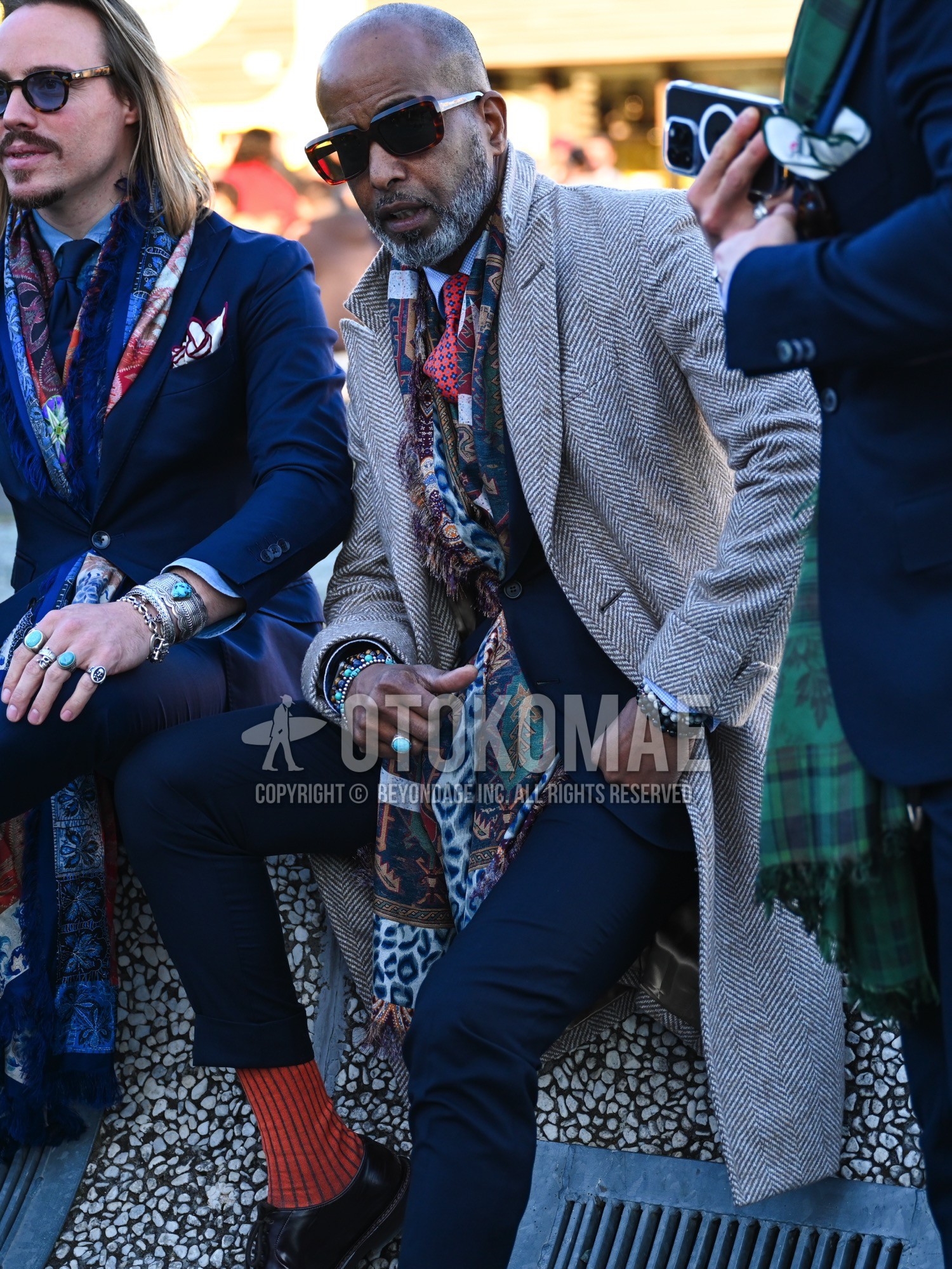 Men's autumn winter outfit with brown tortoiseshell sunglasses, multi-color green red navy yellow graphic bandana/neckerchief, beige brown herringbone ulster coat, white plain shirt, red stripes socks, black plain toe leather shoes, navy plain suit, red small crest necktie.