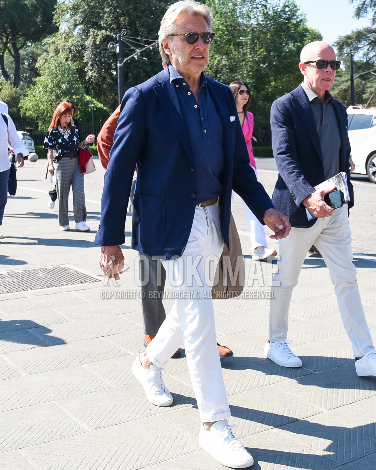 Men's spring summer outfit with brown tortoiseshell sunglasses, navy plain tailored jacket, navy plain polo shirt, brown plain leather belt, white plain cotton pants, white low-cut sneakers.