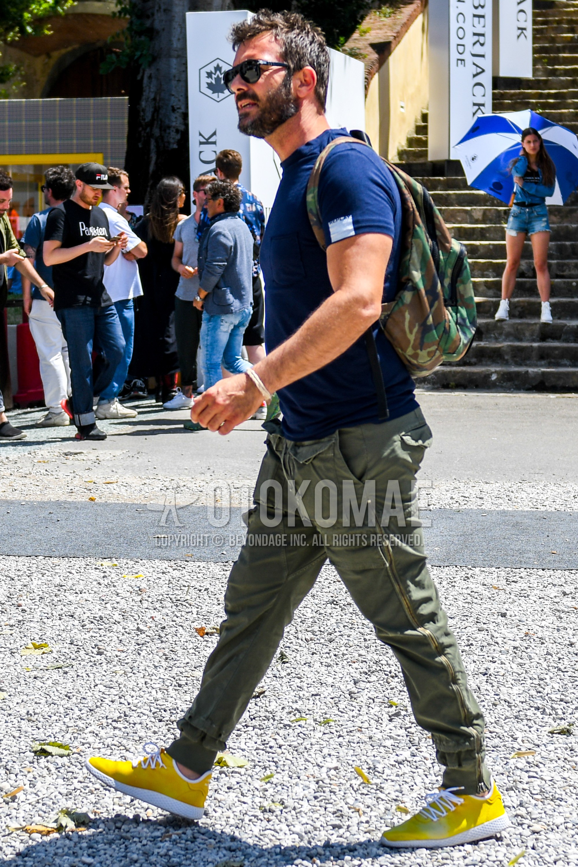 Men's summer outfit with black plain sunglasses, blue plain t-shirt, olive green plain cargo pants, yellow low-cut sneakers, olive green black beige camouflage backpack.