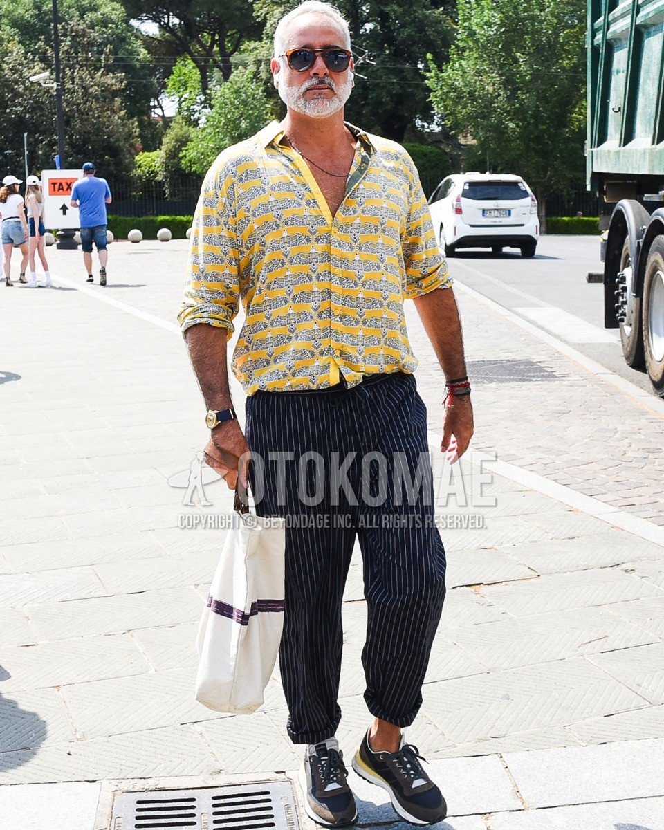 Men's spring summer outfit with pink tortoiseshell sunglasses, yellow graphic shirt, navy stripes easy pants, black low-cut sneakers, white bag tote bag.