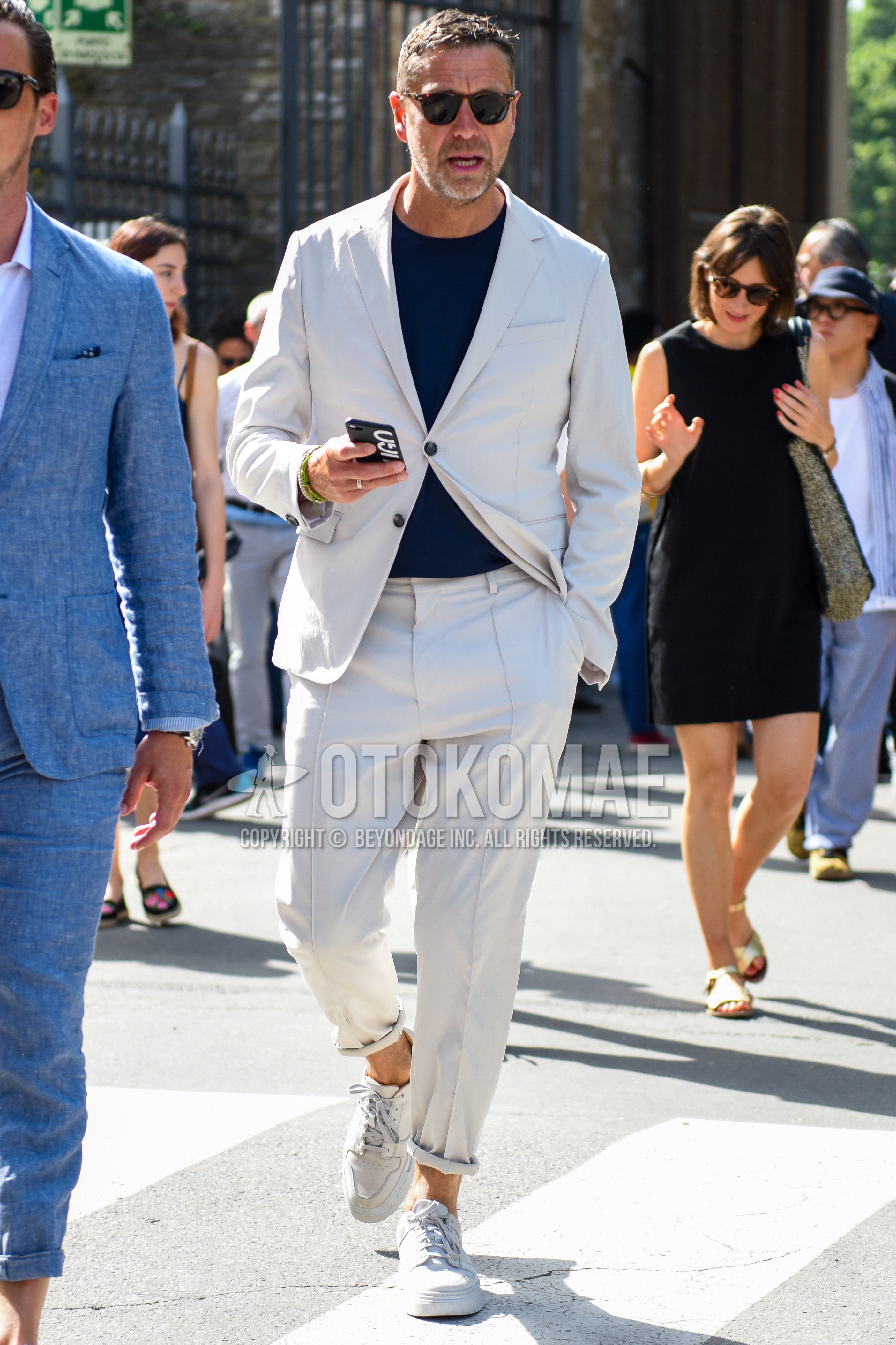 Men's spring summer outfit with brown tortoiseshell sunglasses, navy plain t-shirt, white low-cut sneakers, beige plain suit.