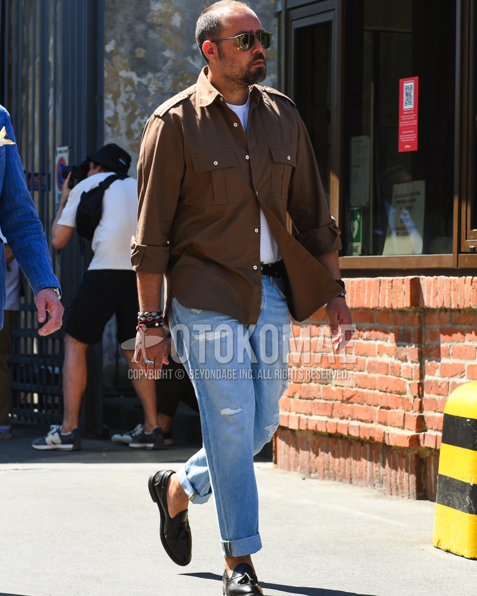 Men's spring summer outfit with olive green plain sunglasses, brown plain military jacket, brown plain shirt jacket, white plain t-shirt, black plain leather belt, blue plain damaged jeans, black tassel loafers leather shoes.