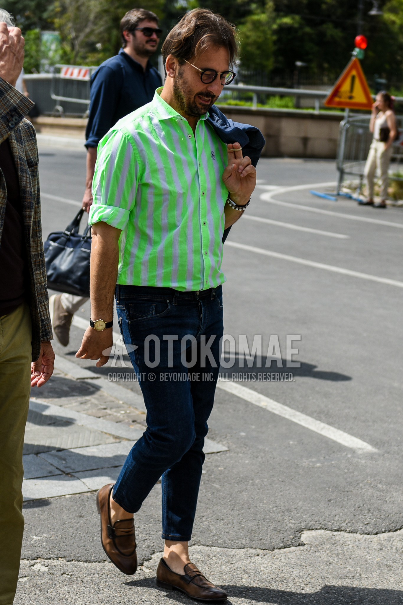 Men's spring summer outfit with black gold plain glasses, green white stripes shirt, navy plain denim/jeans, brown coin loafers leather shoes.