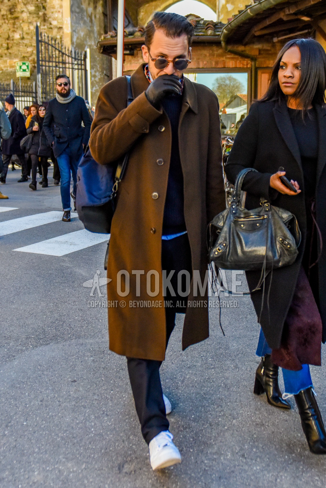 Men's autumn winter outfit with brown tortoiseshell sunglasses, brown plain stenkarrer coat, dark gray plain sweater, gray plain slacks, white low-cut sneakers, black plain backpack.