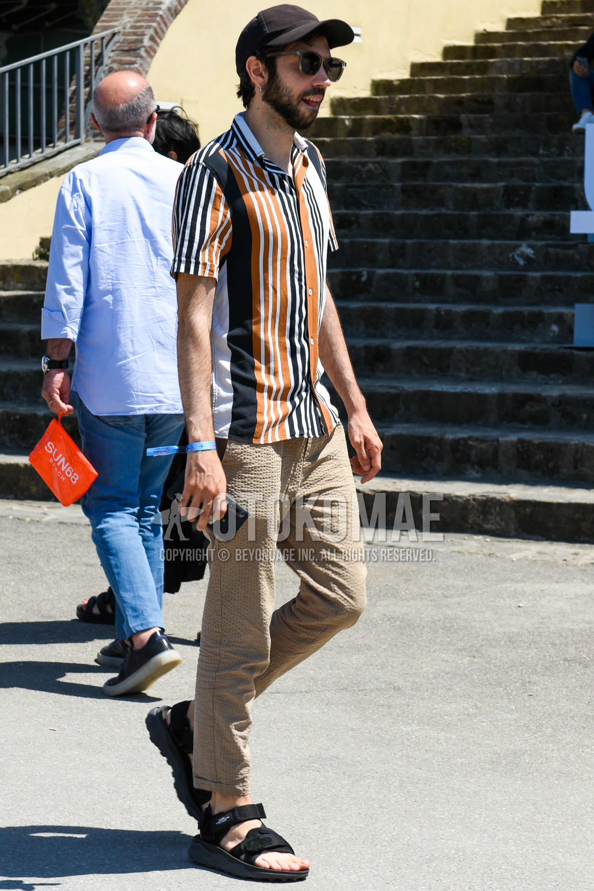 Men's spring summer outfit with black plain baseball cap, black plain sunglasses, multi-color stripes shirt, beige plain chinos, black sport sandals.