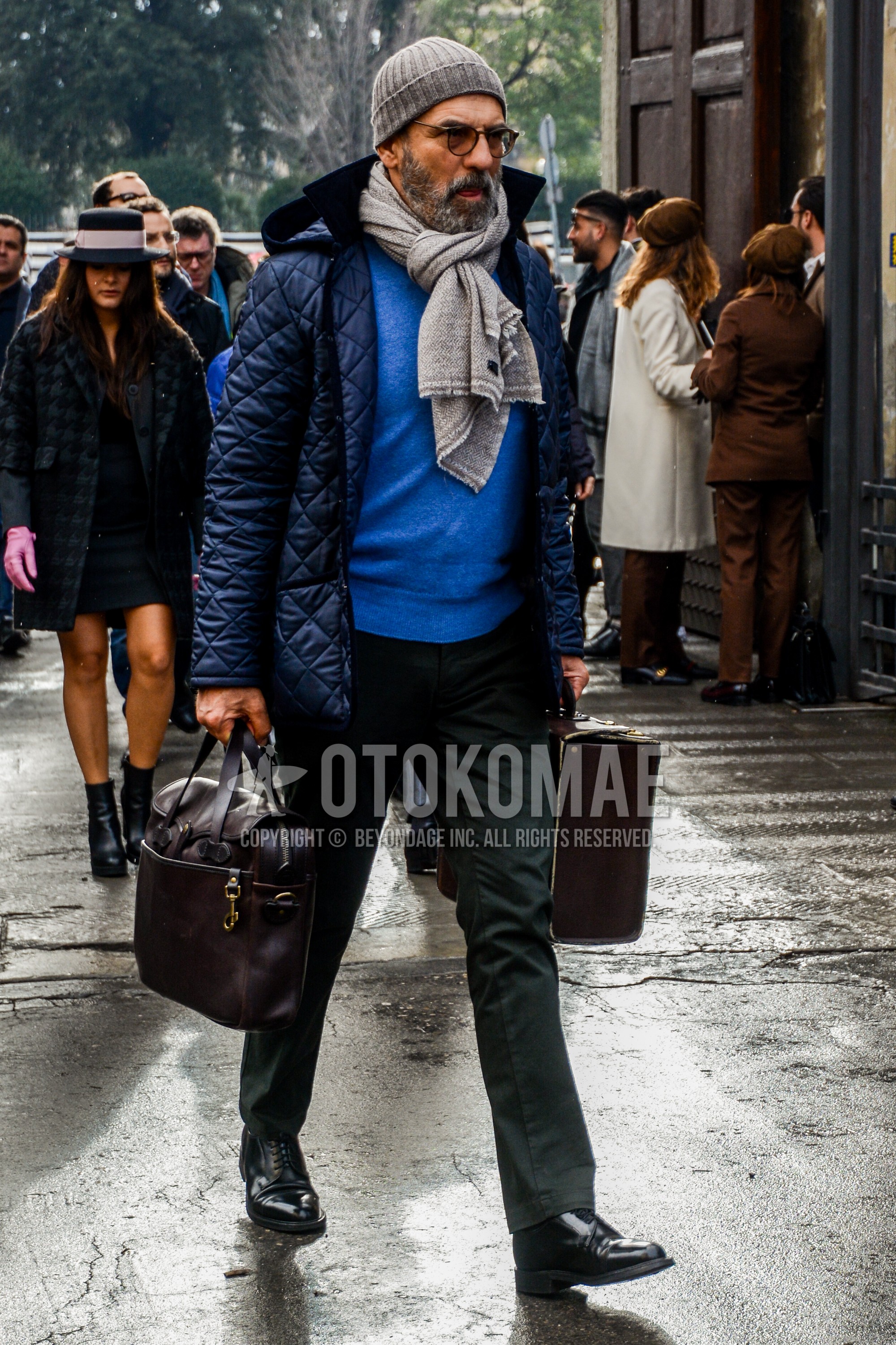 Men's autumn winter outfit with plain knit cap, plain glasses, beige plain scarf, navy plain quilted jacket, blue plain sweater, gray plain slacks, black plain toe leather shoes, brown plain briefcase/handbag.