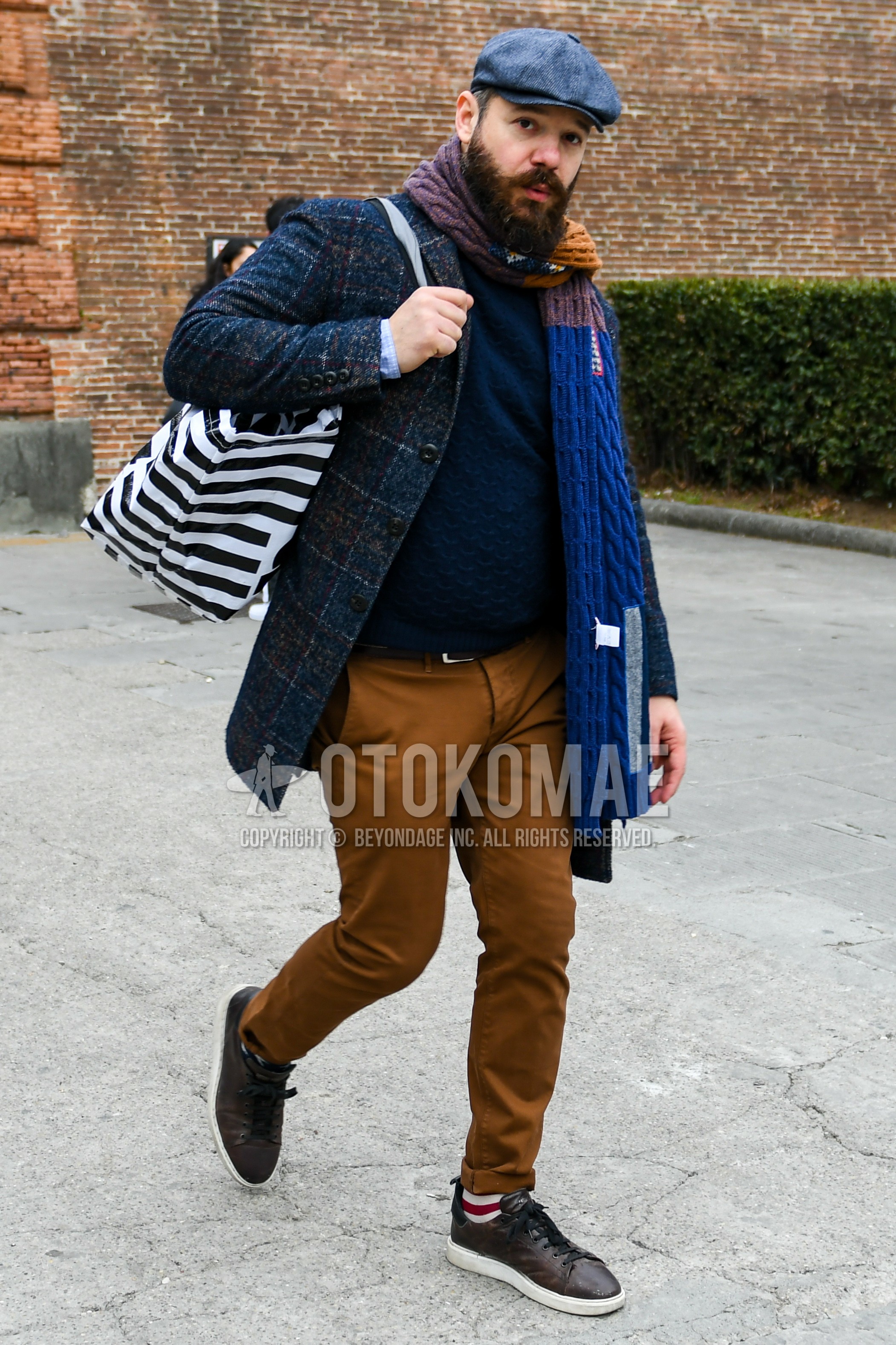 Men's autumn winter outfit with gray plain cap, blue beige brown check scarf, gray check chester coat, gray plain sweater, plain leather belt, brown plain chinos, white red horizontal stripes socks, brown low-cut sneakers, white black horizontal stripes tote bag.
