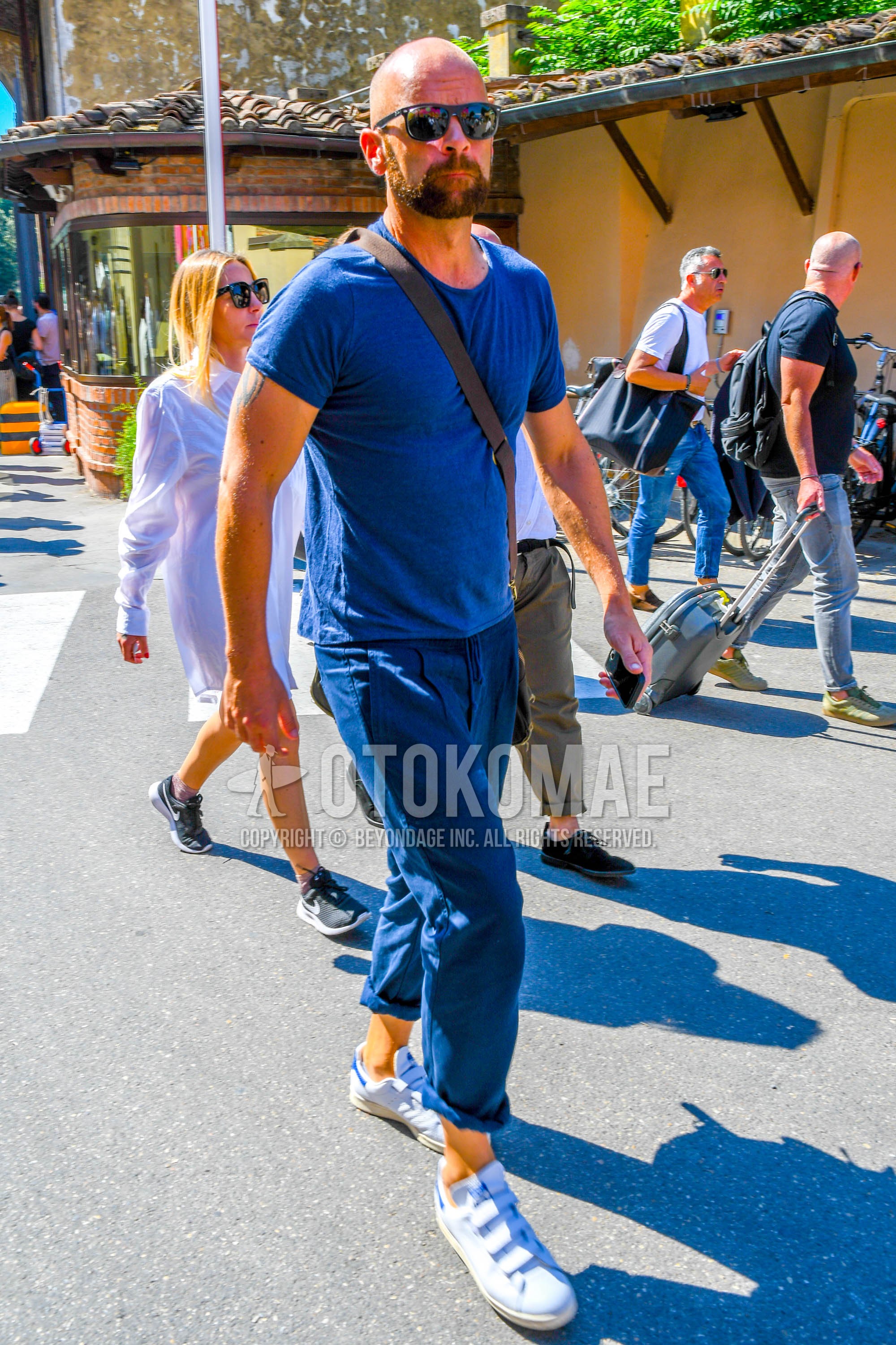 Men's summer outfit with plain sunglasses, blue plain t-shirt, blue plain pleated pants, white low-cut sneakers.