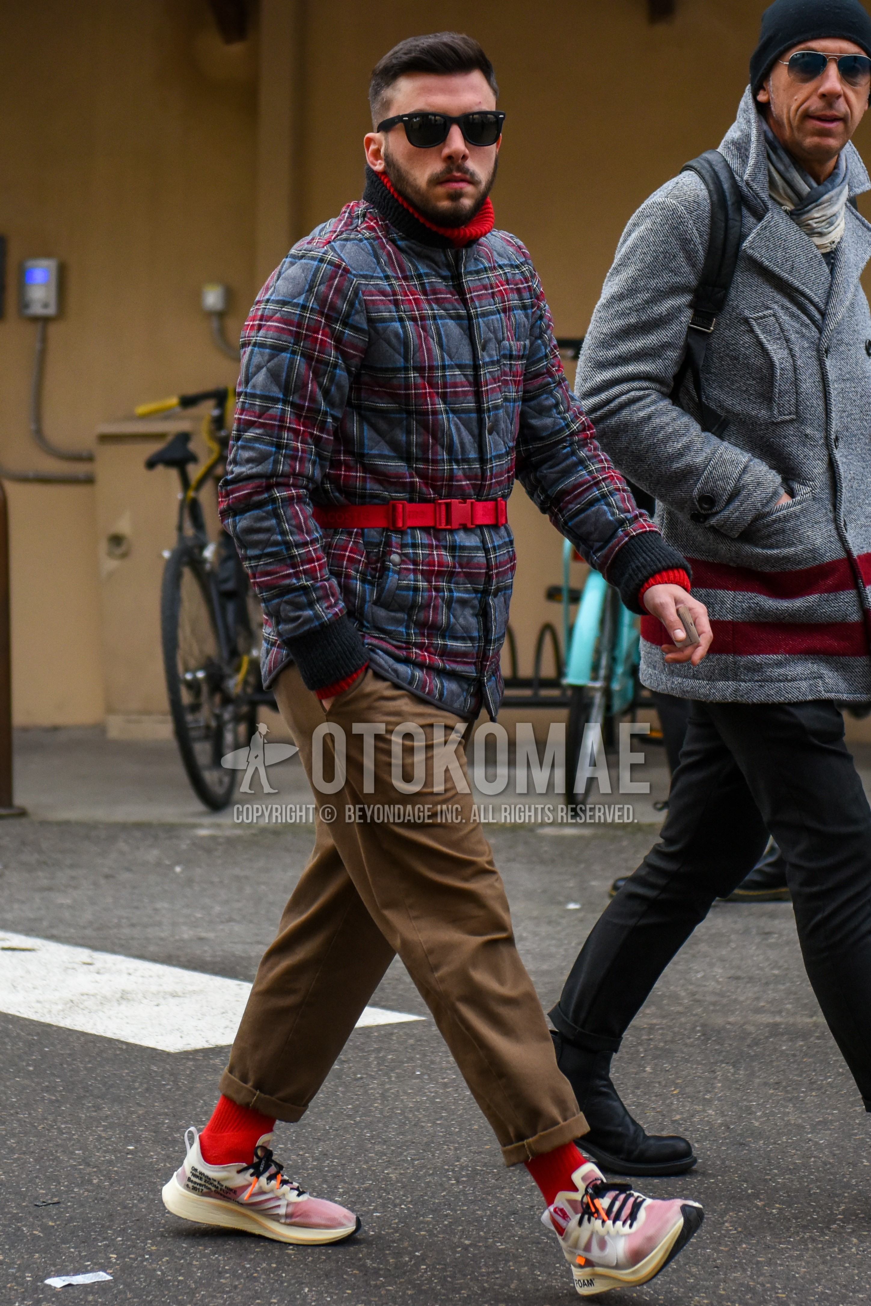 Men's winter outfit with black plain sunglasses, gray red check quilted jacket, brown plain chinos, red plain socks, white low-cut sneakers.