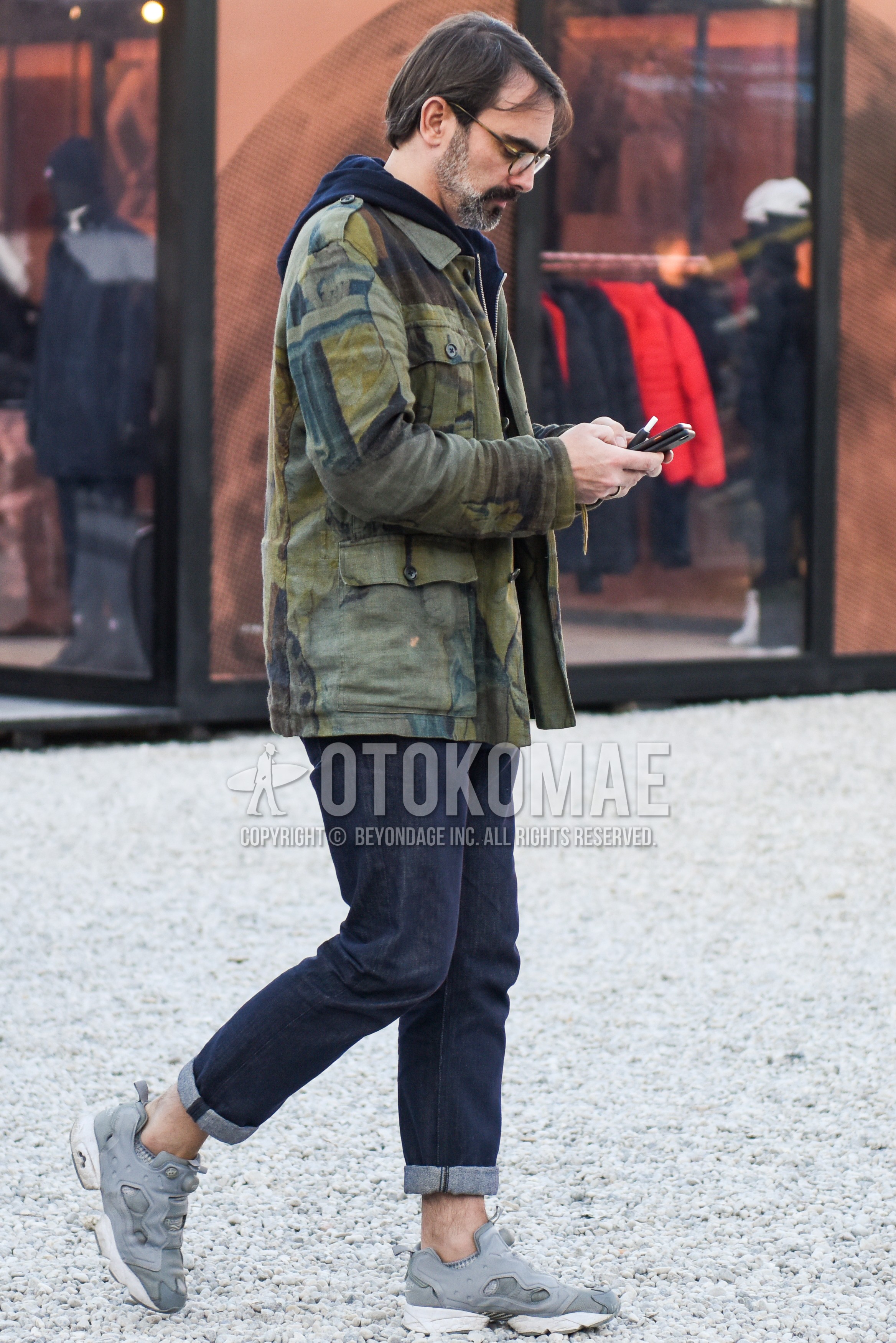 Men's autumn winter outfit with brown tortoiseshell glasses, olive green outerwear shirt jacket, navy plain hoodie, navy plain denim/jeans, gray low-cut sneakers.