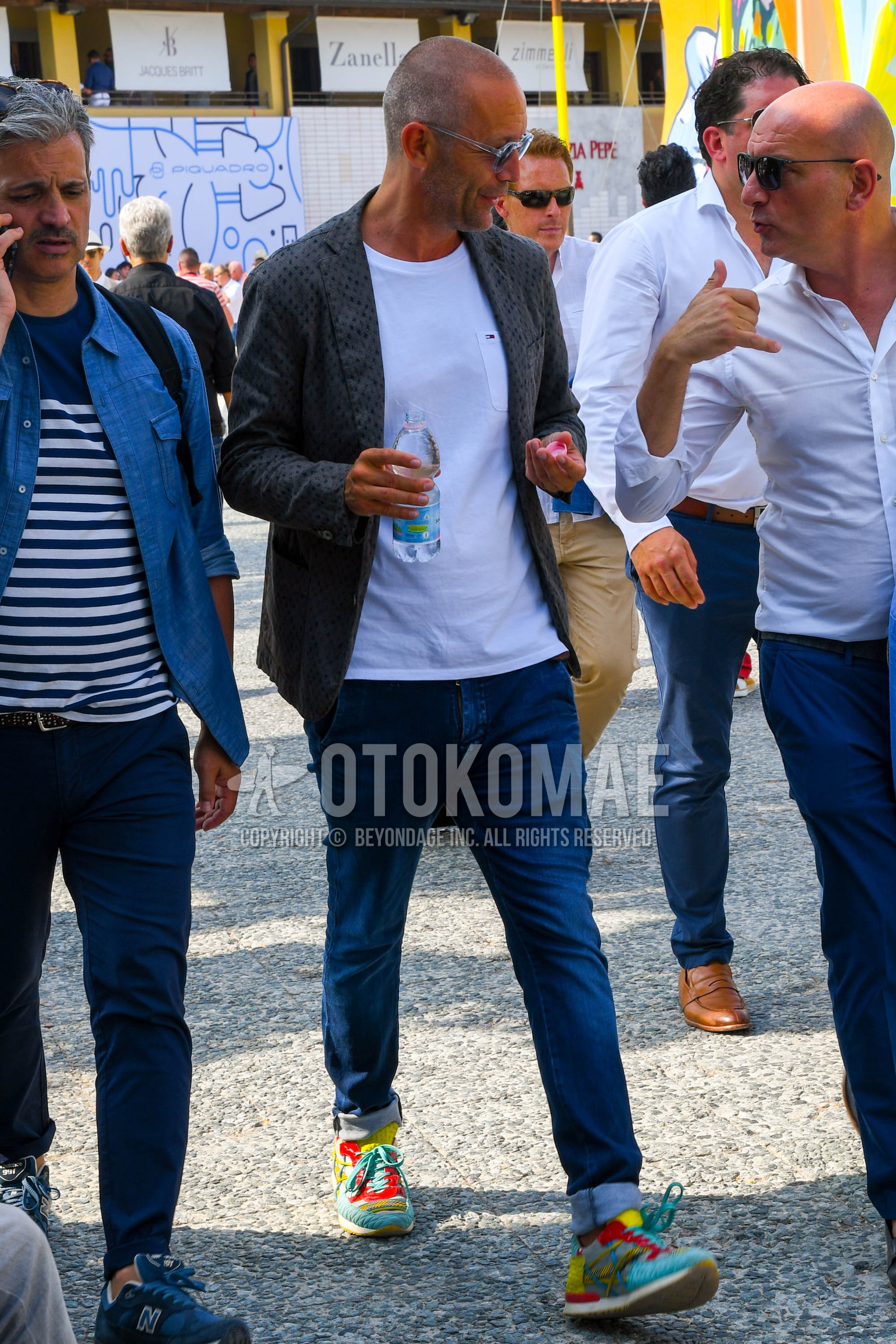 Men's spring summer autumn outfit with clear plain sunglasses, gray outerwear tailored jacket, white plain t-shirt, blue plain denim/jeans, multi-color low-cut sneakers.
