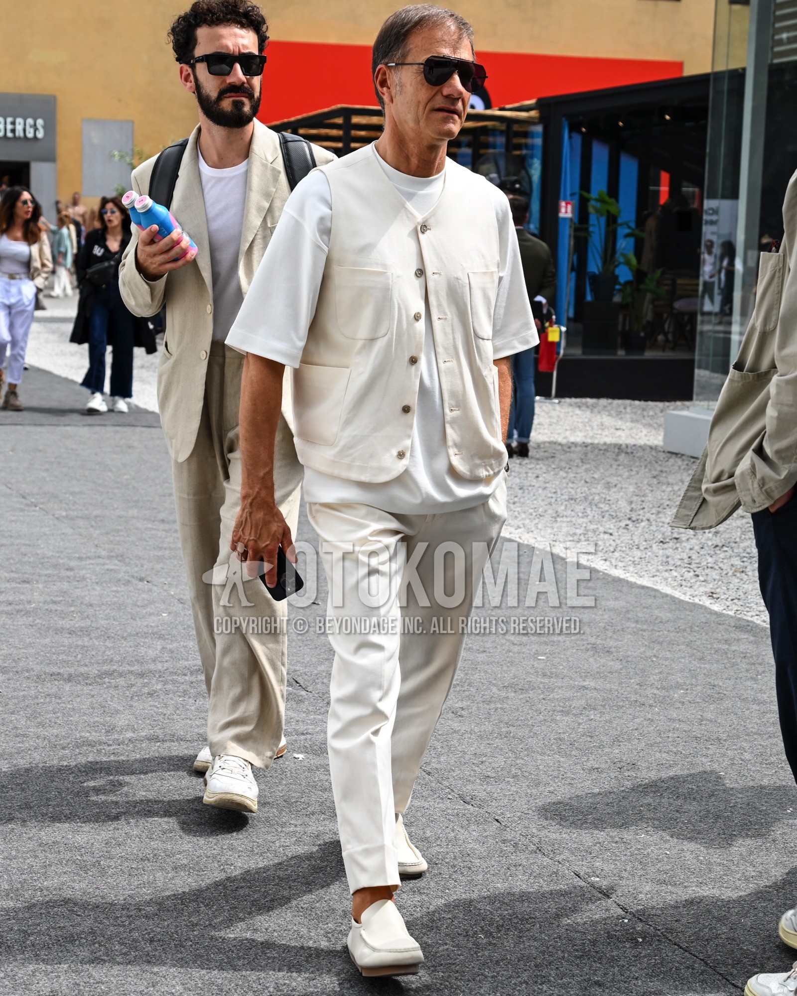 Men's spring summer outfit with black plain sunglasses, white plain casual vest, white plain t-shirt, white plain slacks, white slip-on sneakers.