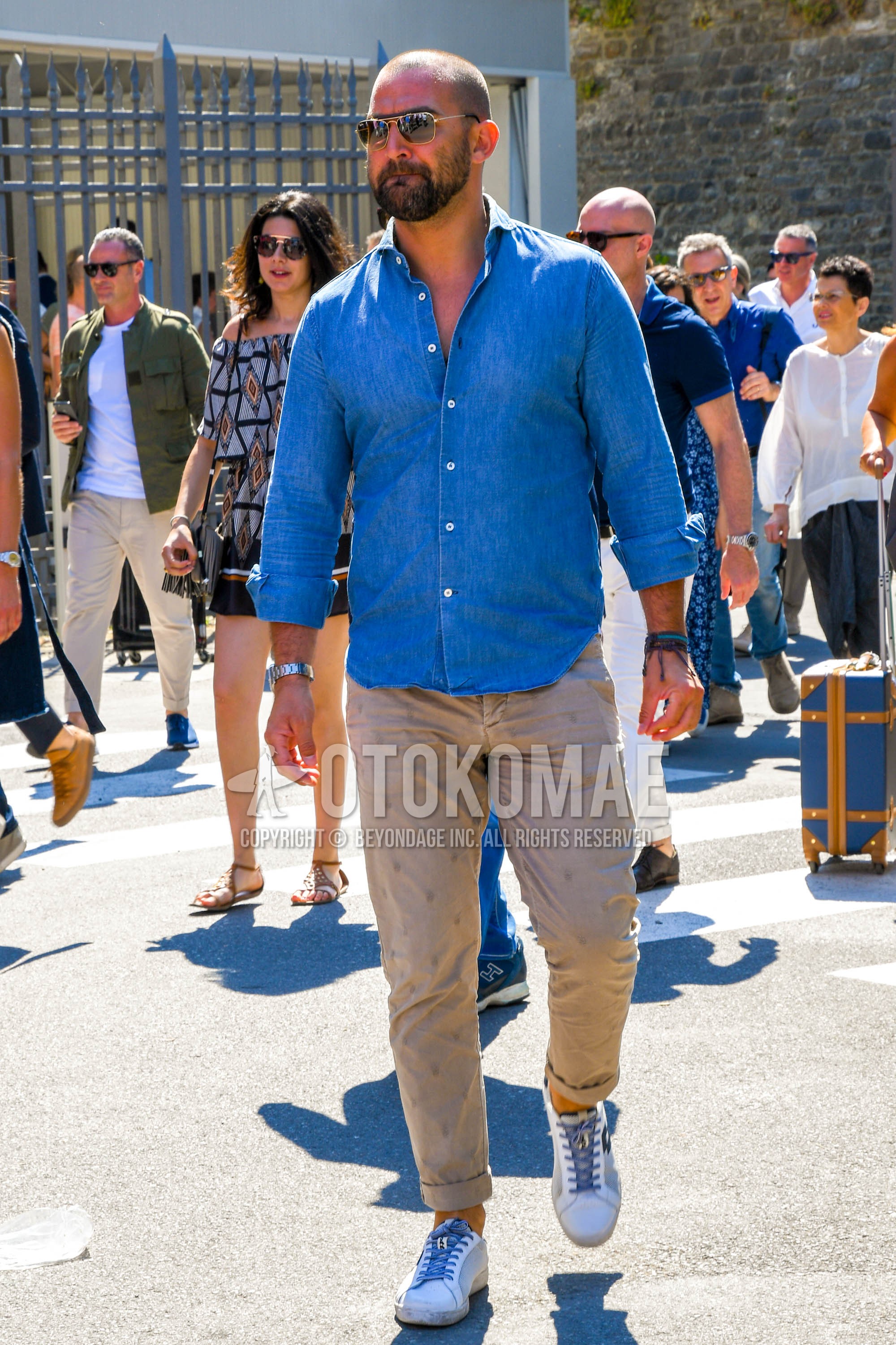 Men's spring summer outfit with plain sunglasses, blue plain denim shirt/chambray shirt, beige bottoms chinos, white low-cut sneakers.