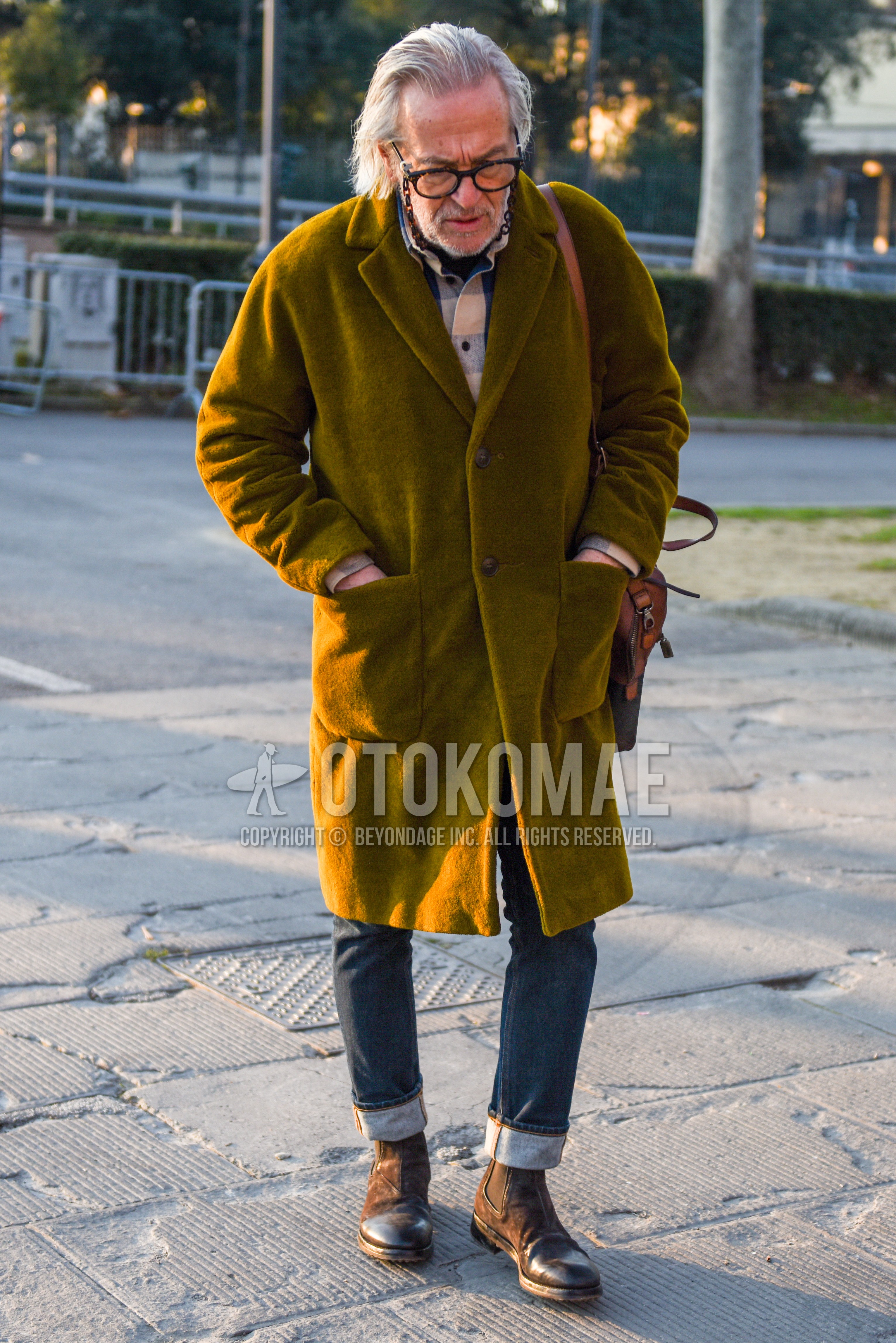 Men's autumn winter outfit with beige tortoiseshell glasses, olive green plain chester coat, multi-color check shirt, navy plain denim/jeans, brown side-gore boots.