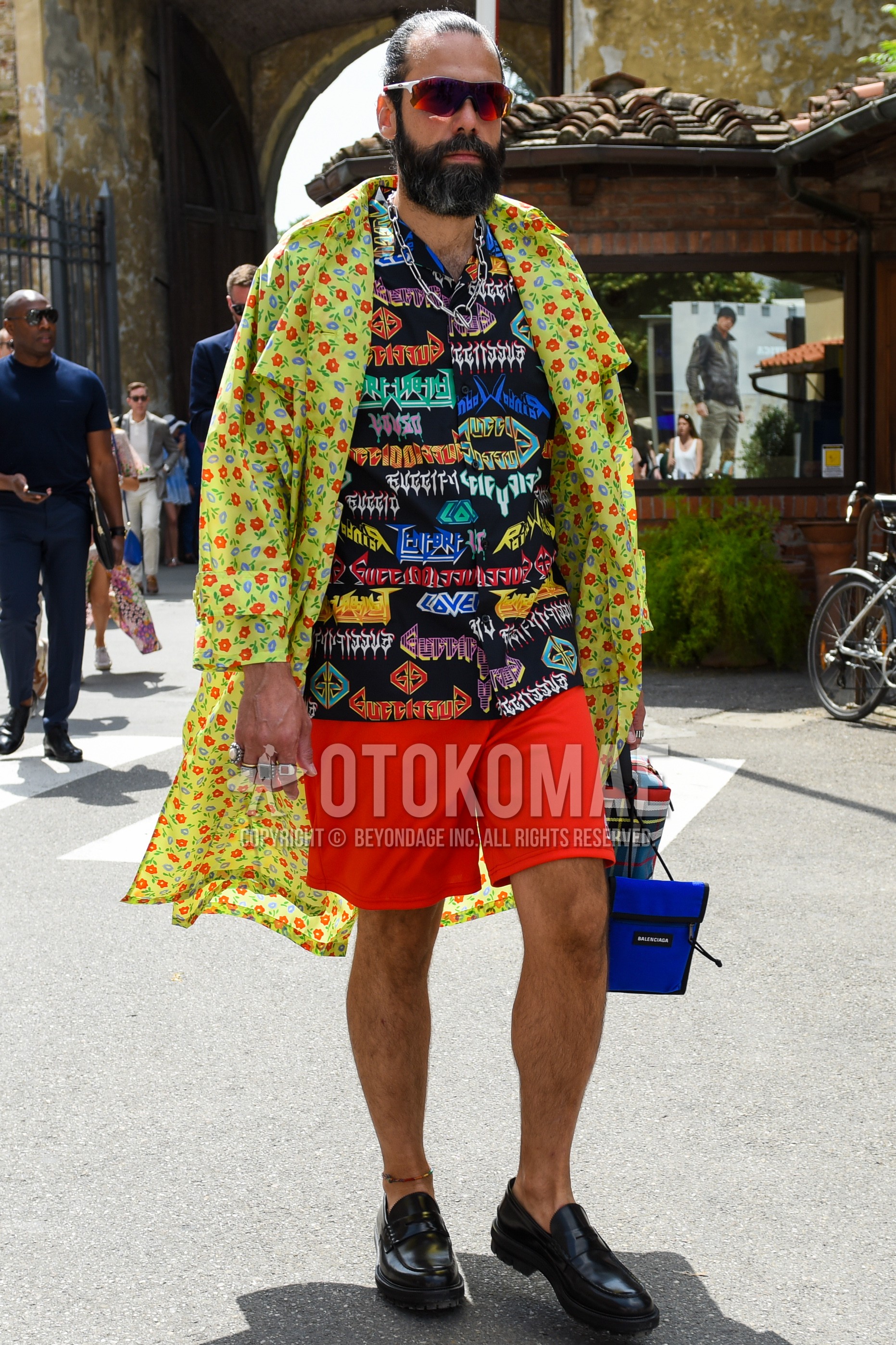 Men's spring summer autumn outfit with white red plain sunglasses, yellow botanical trench coat, multi-color graphic shirt, red plain short pants, black coin loafers leather shoes.