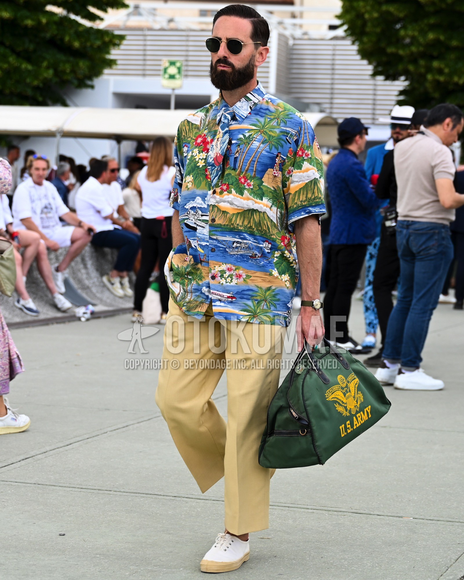 Men's spring summer outfit with black plain sunglasses, blue whole pattern shirt, beige plain chinos, white low-cut sneakers, olive green deca logo boston bag.