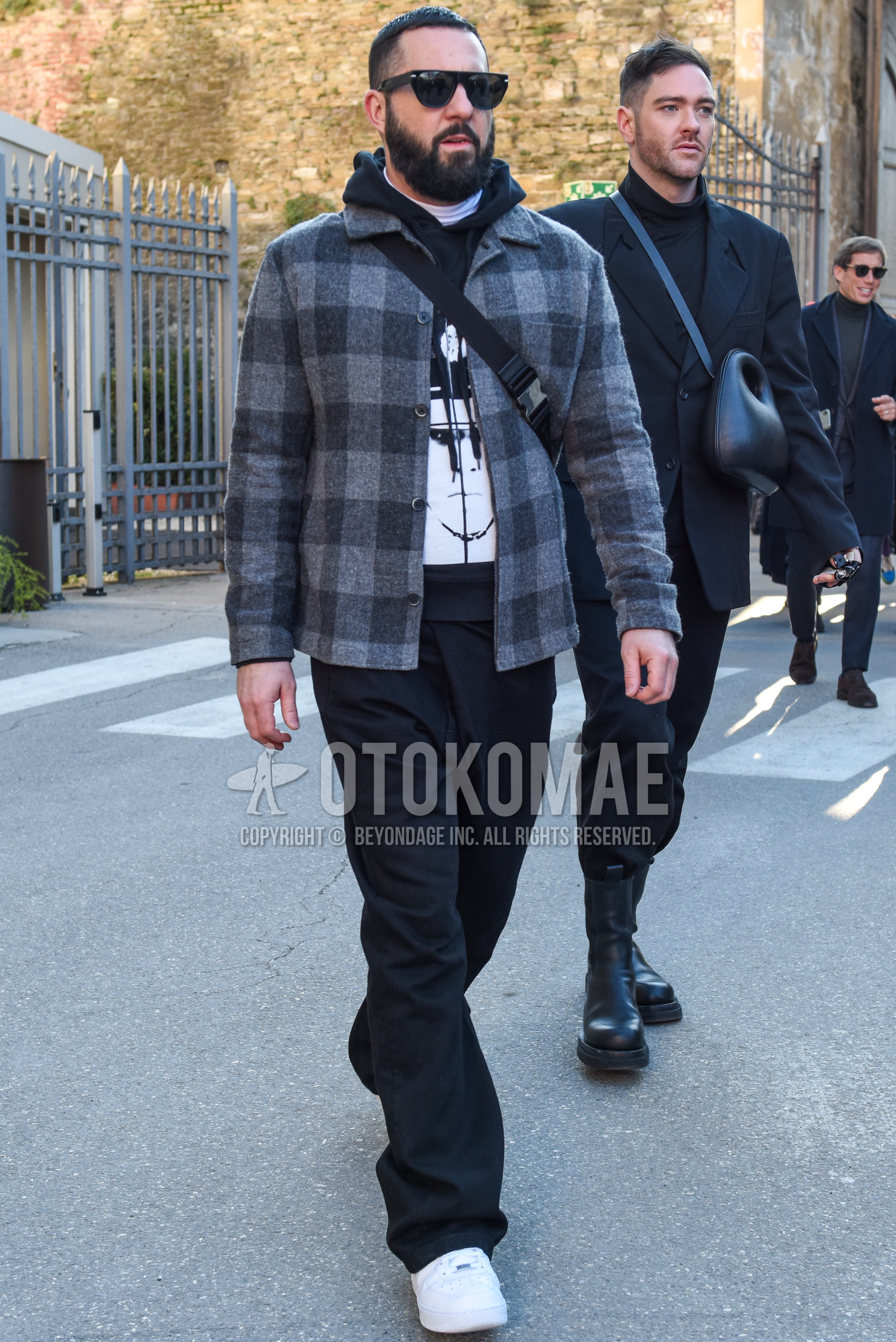 Men's autumn winter outfit with black plain sunglasses, gray black check shirt jacket, black graphic hoodie, dark gray plain slacks, white low-cut sneakers.