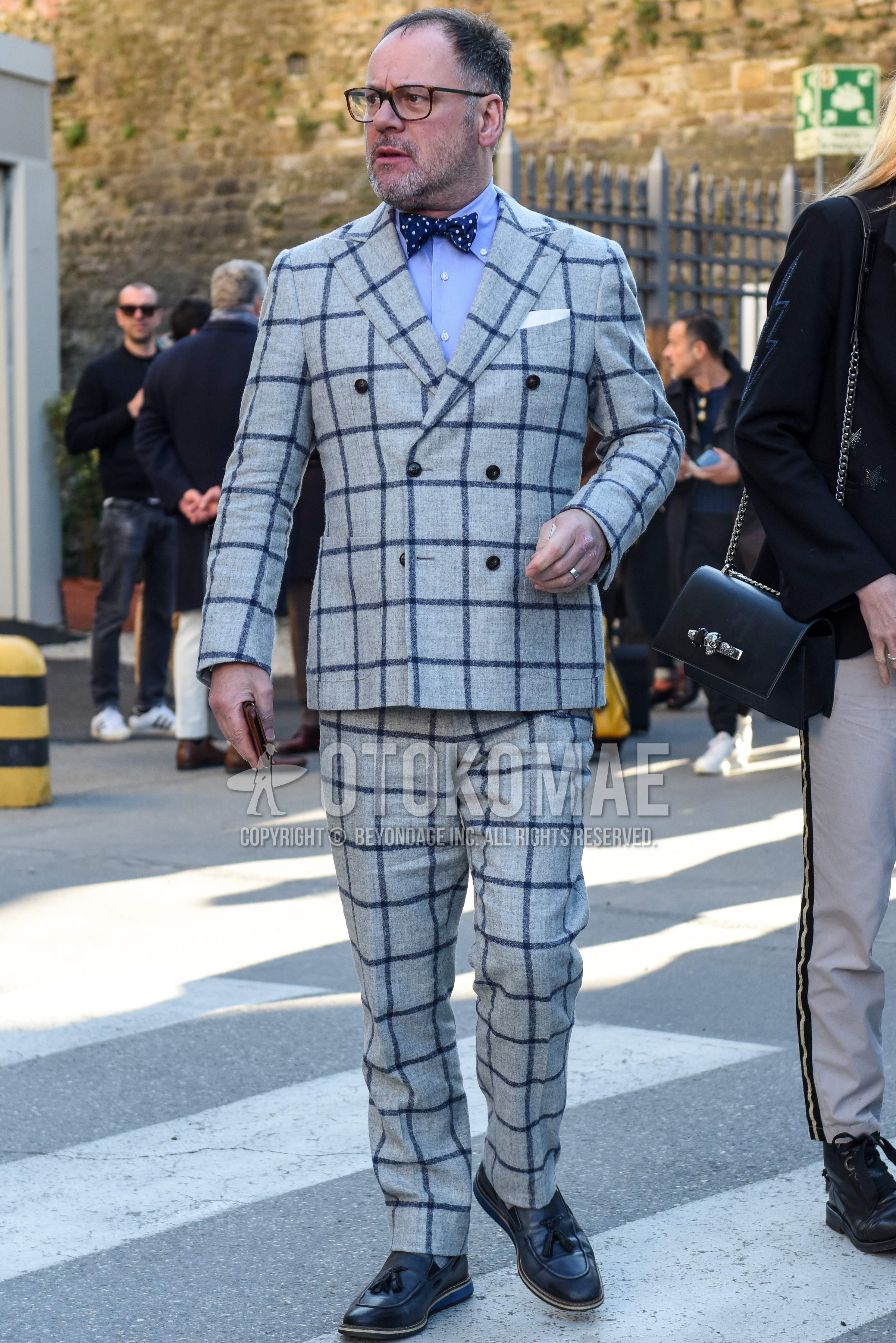 Men's spring autumn outfit with brown tortoiseshell glasses, light blue plain shirt, black tassel loafers leather shoes, gray check suit, navy dots bow tie.