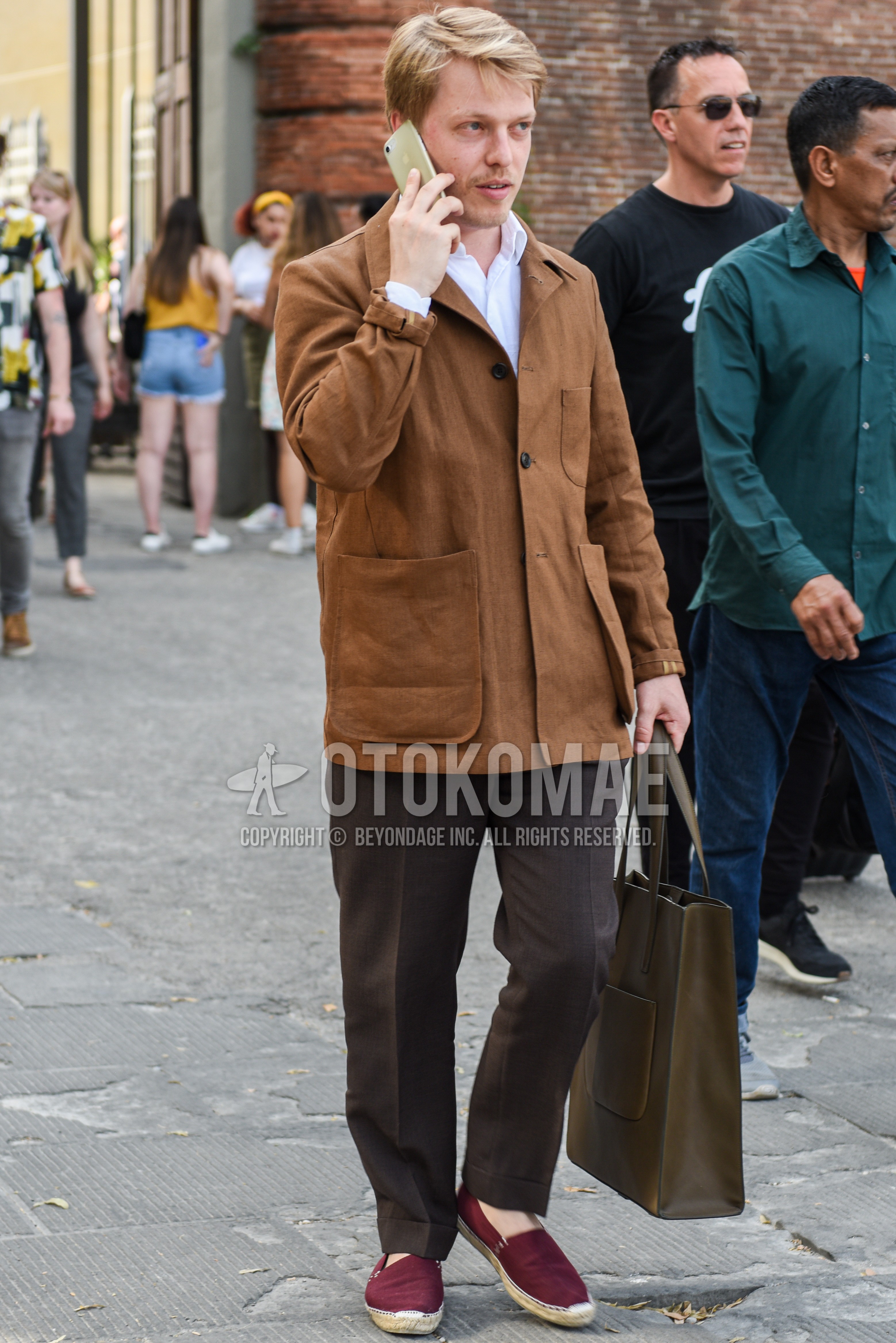 Men's spring autumn outfit with brown plain shirt jacket, white plain shirt, brown plain slacks, red slip-on sneakers, olive green plain tote bag.