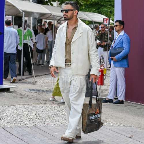 ウェリントンの黒無地のサングラス、ブラウン無地のシャツ、白ストライプのシャツ、白・黒ベルトのメッシュベルト、白・黒ベルトのレザーベルト、白ストライプのスラックス、白ローファーの革靴、マルチカラーレタードのトートバッグ、白ストライプのカジュアルセットアップを合わせた春夏のメンズコーデ・着こなし。